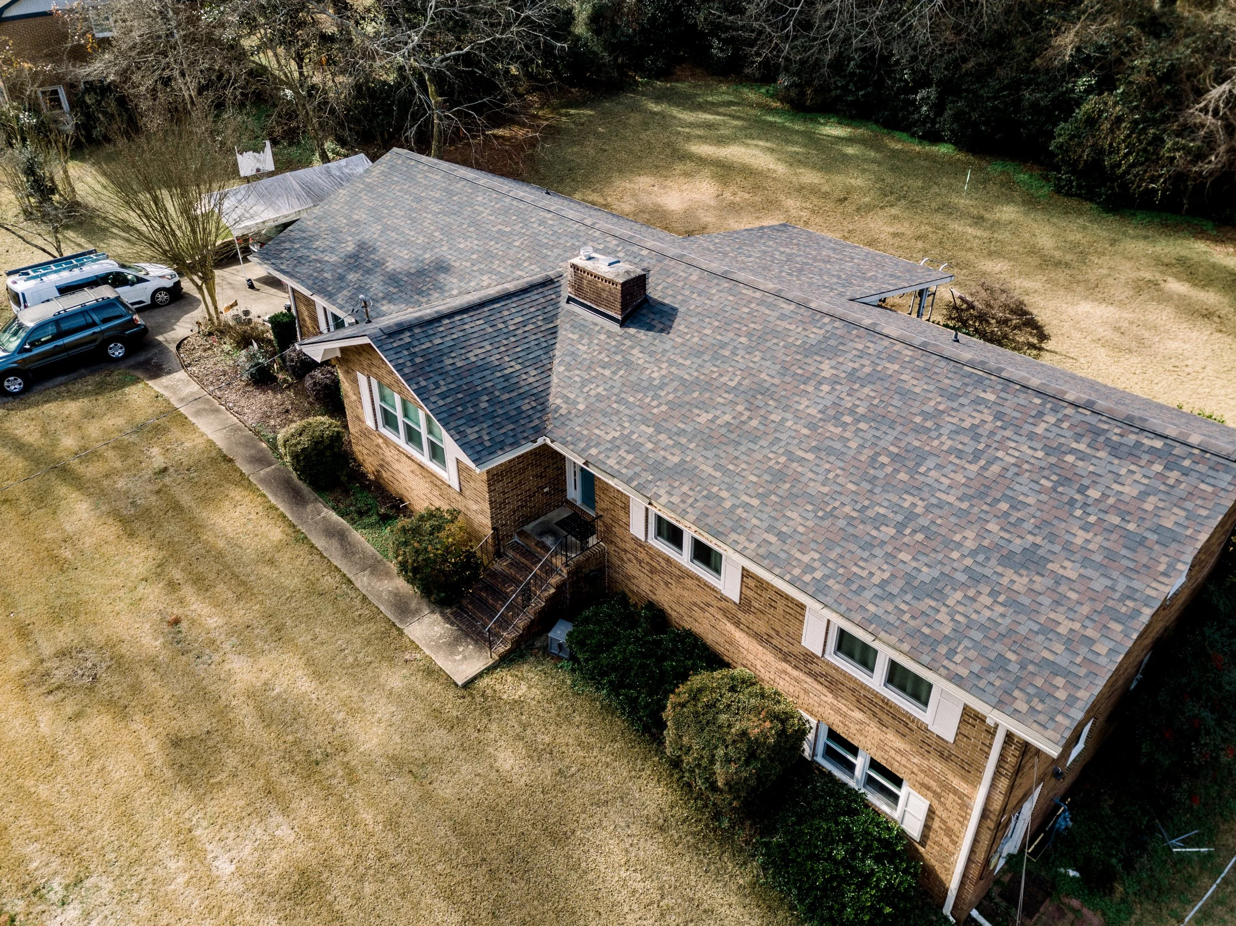 An aerial view of a brick house with a gray shingled roof, surrounded by a yard with grass, bushes, and trees. Cars are parked on the driveway and a lawn.