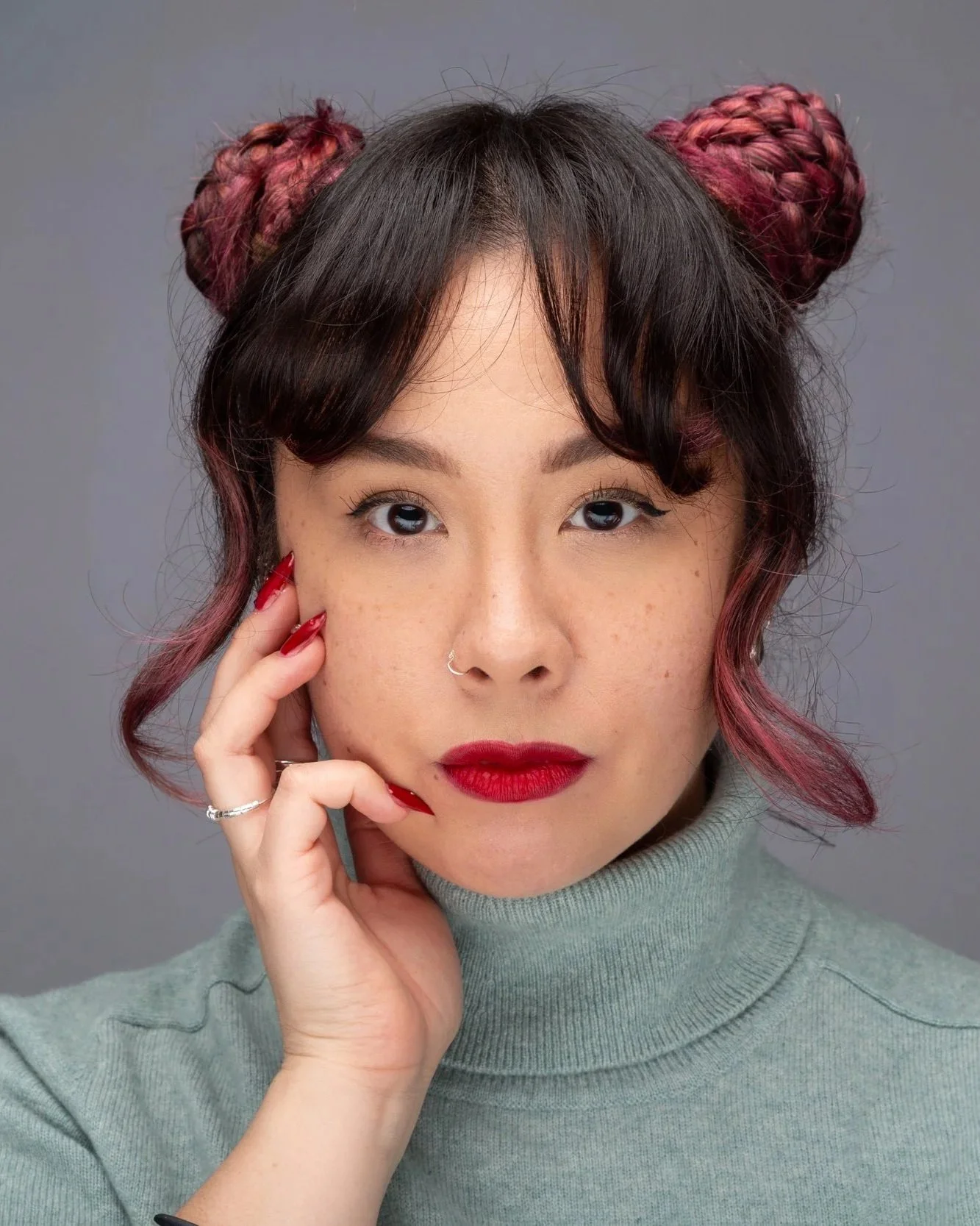 Close-up of a woman with dark hair styled in buns with pink highlights, wearing a gray turtleneck sweater and red lipstick, touching her face with her hand.