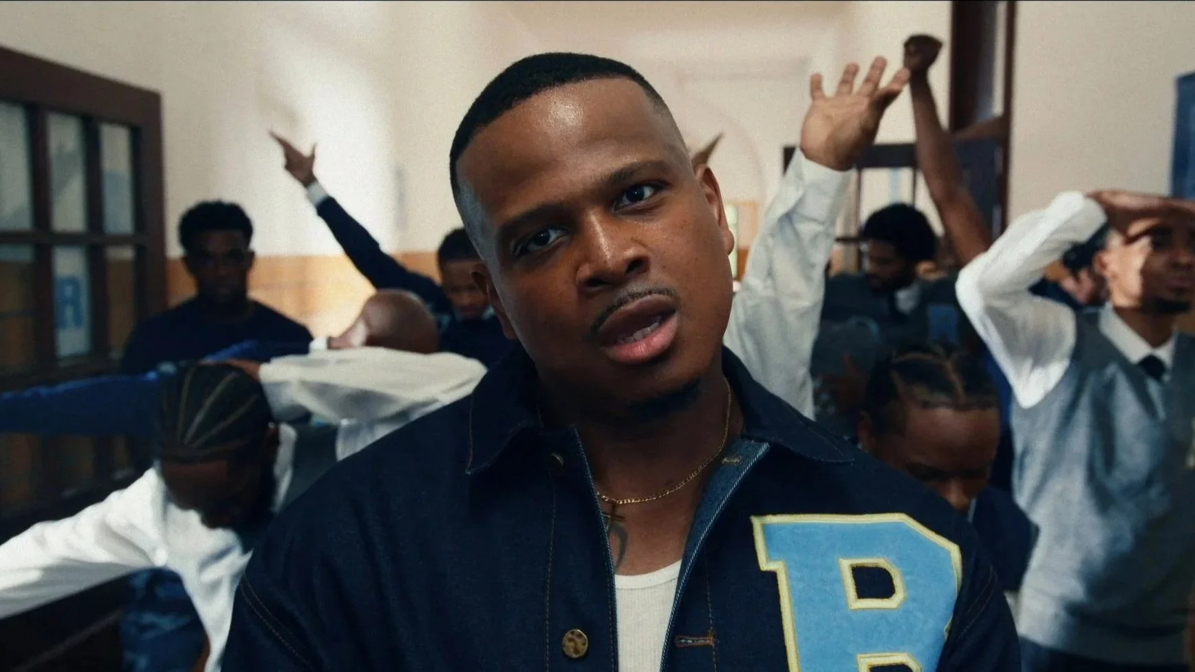 A young man with short hair and a chain necklace looks directly at the camera in a classroom full of students, some raising their hands, with the focus on him.