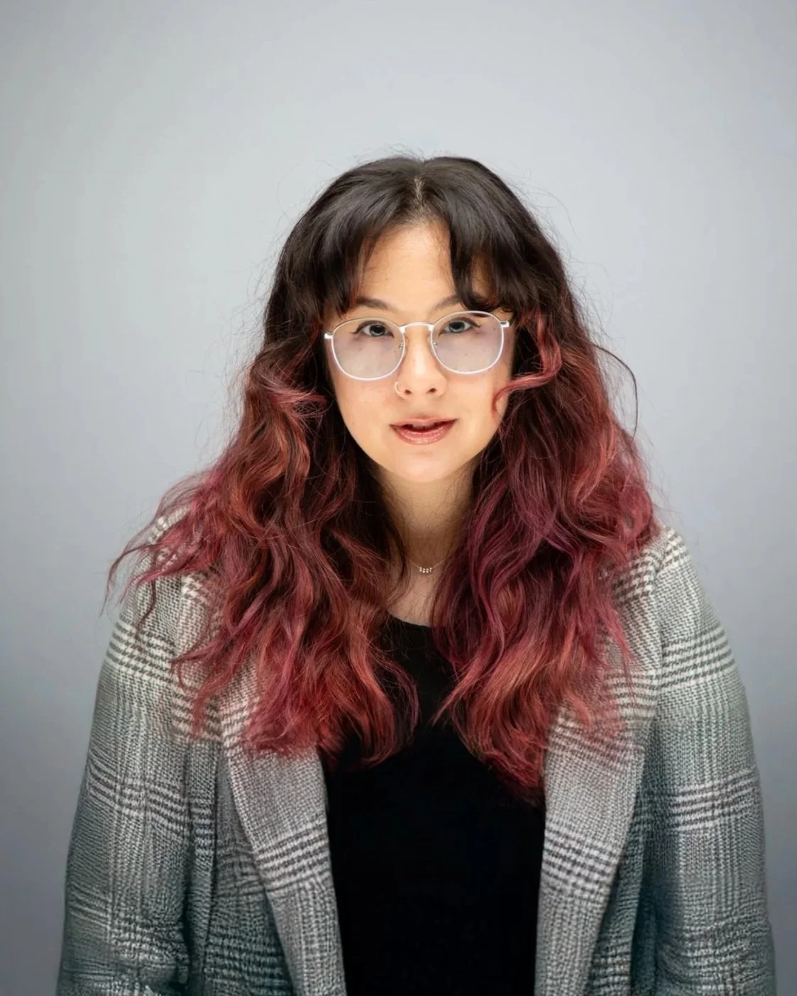 A woman with glasses and wavy hair that is dark at the top and red at the ends. She is wearing a plaid blazer over a black top, with a plain light gray background behind her.