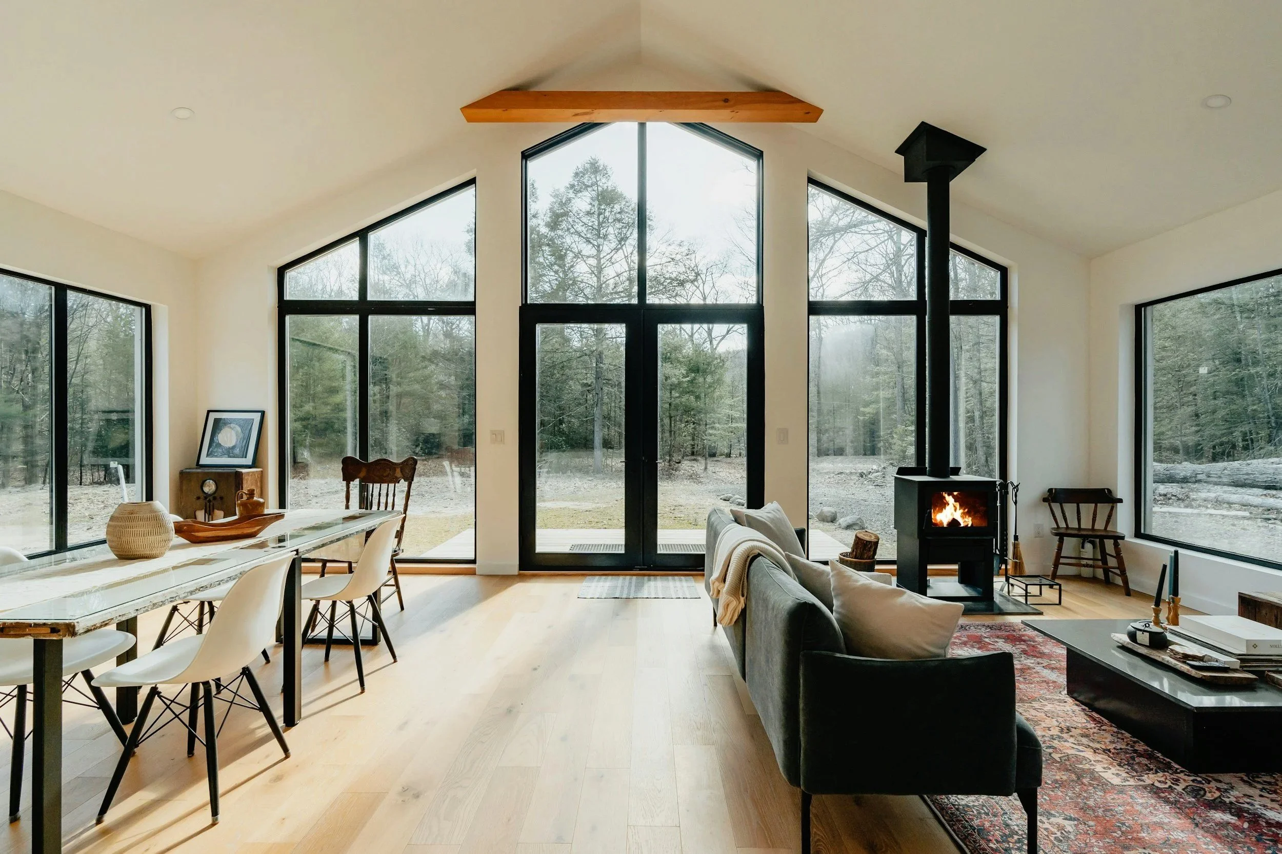 Spacious living room with large floor-to-ceiling windows, a fireplace, and modern furniture in a home surrounded by woods.