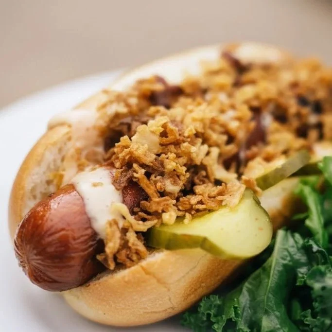 Close-up of a hot dog with chili, shredded cheese, pickles, and lettuce on a bun, served on a white plate.