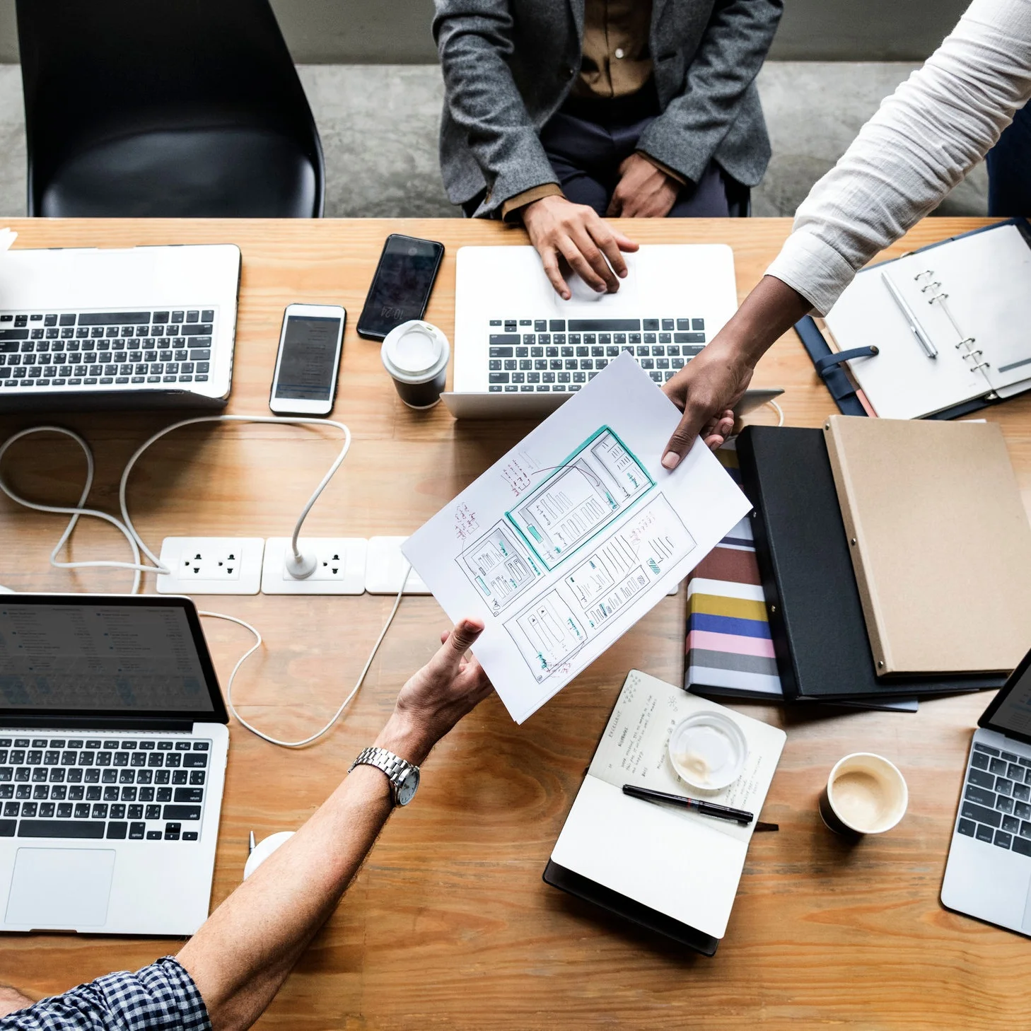 Aerial view of a desk with multiple laptops and people passing documents and making notes. They are hard at work. There are cups of coffee on the desk.
