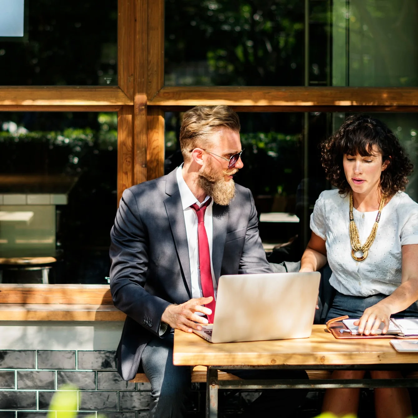A man and woman sitting outside a cafe in discussion, looking at a laptop.
