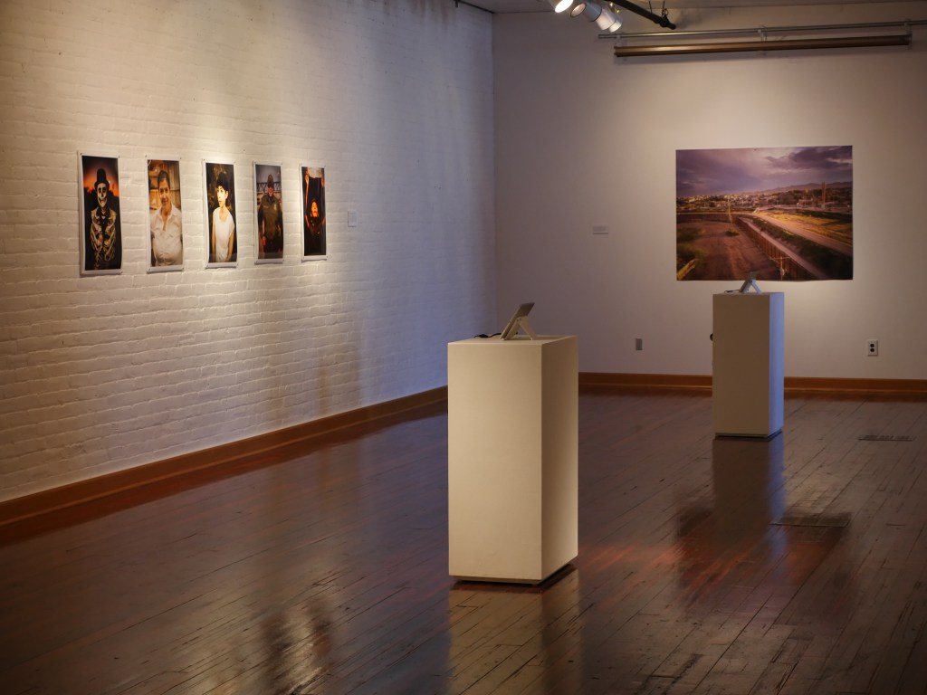 An installation shot of the exhibition shows portraits of El Paso and Juarez locals, a drone shot of the landscape along the fence, and two iPads with videos playing. Image courtesy of the artists.