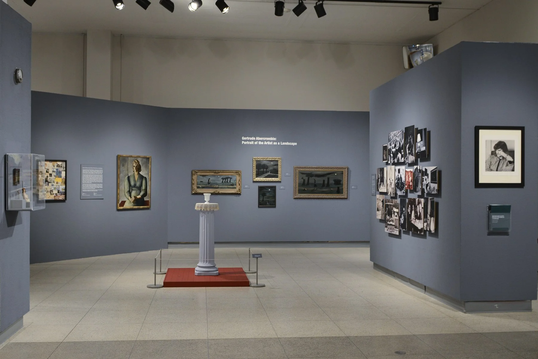 The entrance to the Abercrombie exhibition with dark gray-blue walls, a photo of her hangs to the right, on the back wall are several of her paintings and in the center is the still life display mentioned above. Image courtesy of the Illinois State Museum.
