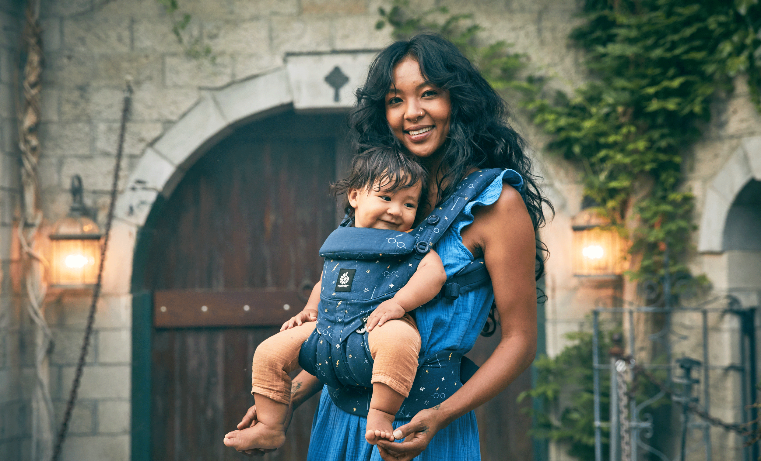 A woman with long dark wavy hair smiling while holding a young toddler in a blue child carrier backpack. The toddler, also smiling, has short dark hair and is wearing a beige outfit. They are outdoors in front of a stone and wooden gate, with greenery and warm lights.