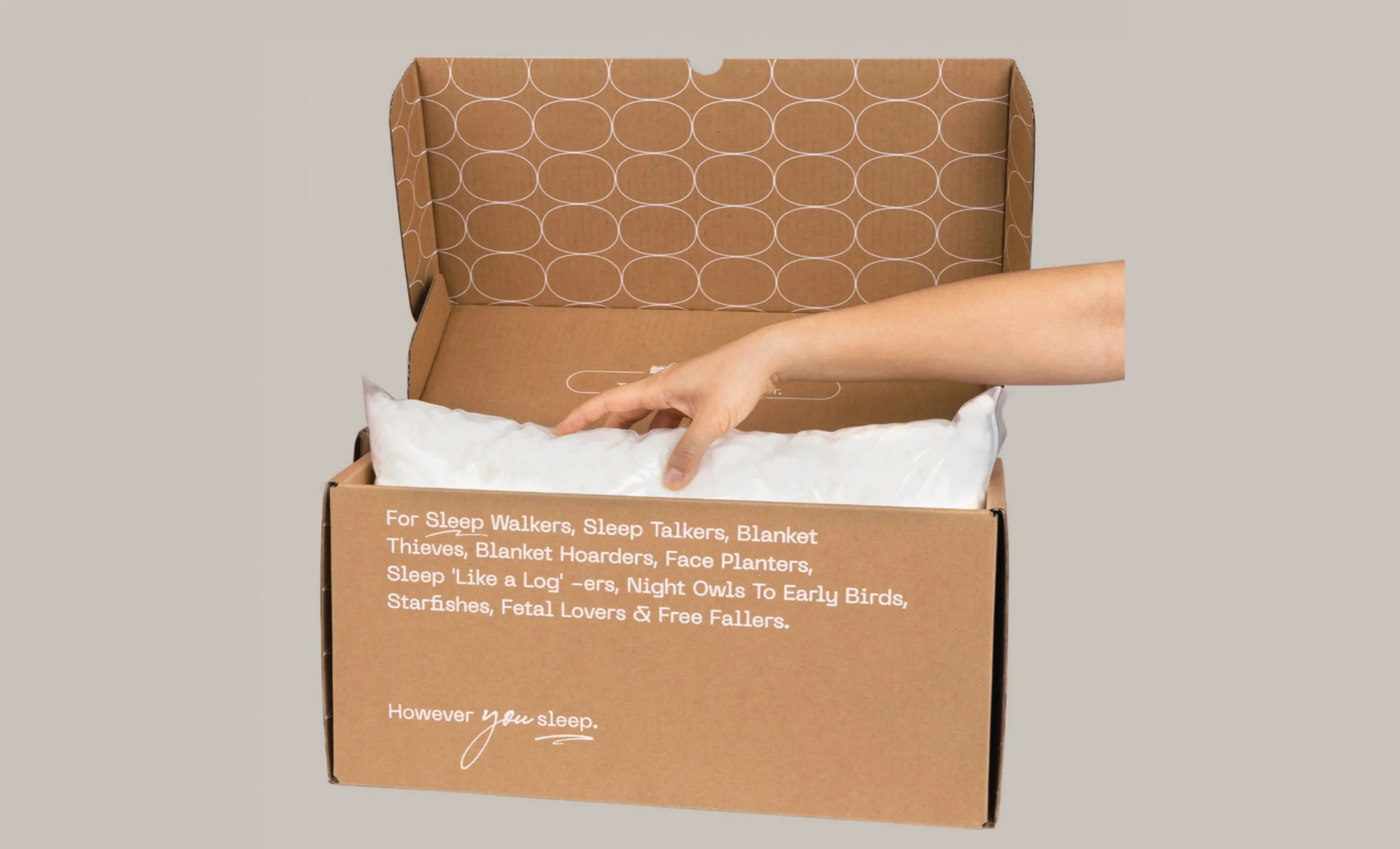 Person's hand placing a white pillow into an open cardboard box with printed text, on a plain gray background.