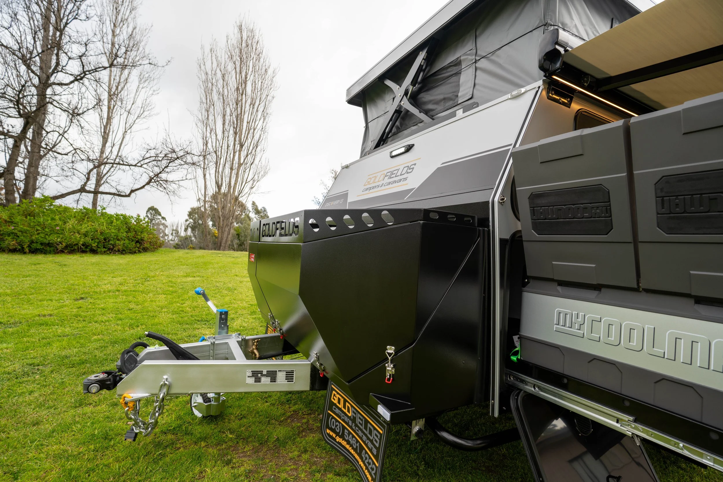Grey camper trailer with black storage boxes attached, parked on a grassy area with leafless trees in the background.