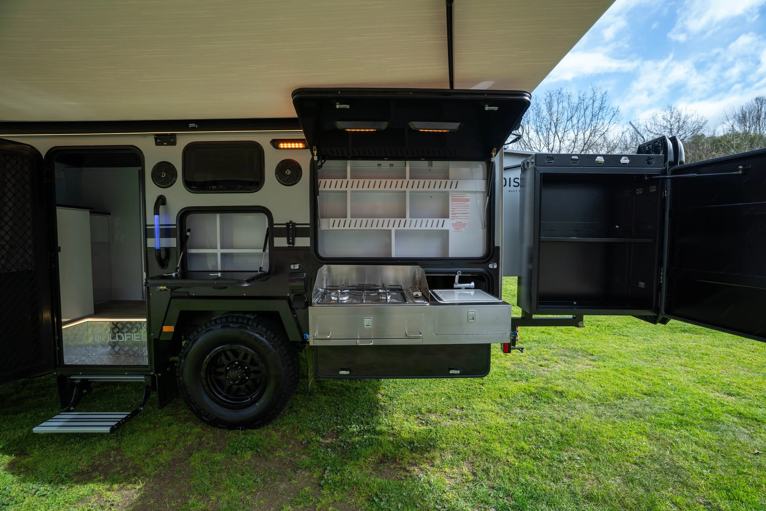 A travel trailer with an open side kitchen, including a stove and storage compartments, parked on grass with trees and sky in the background.