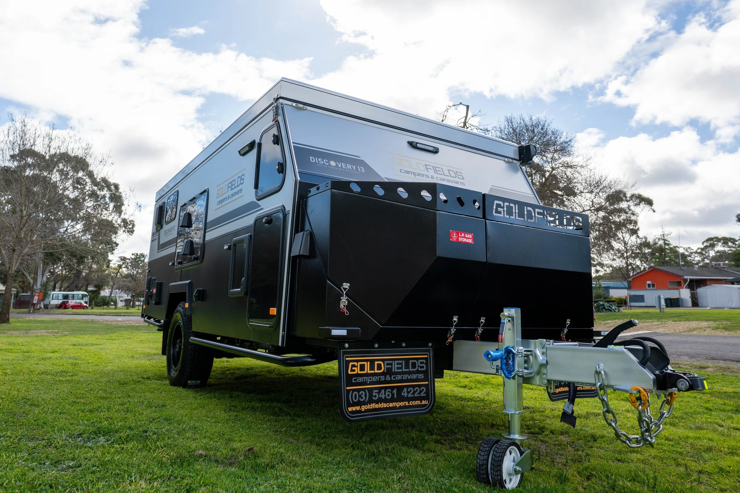A black camper trailer with branding for Gold Fields campers & caravans is parked on grass under a partly cloudy sky. The trailer has a metal frame and hitch, with a sign displaying contact information in front.