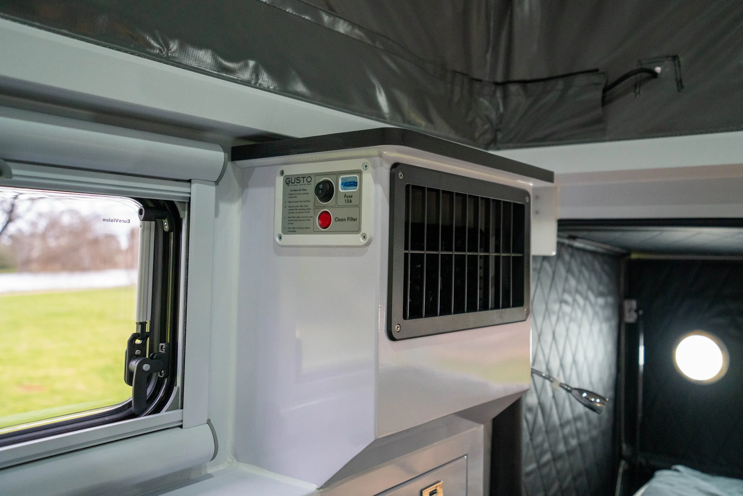 Close-up of a vending machine or food service unit with a control panel, air vent, and part of a window, with blurred background of a grassy outdoor area.