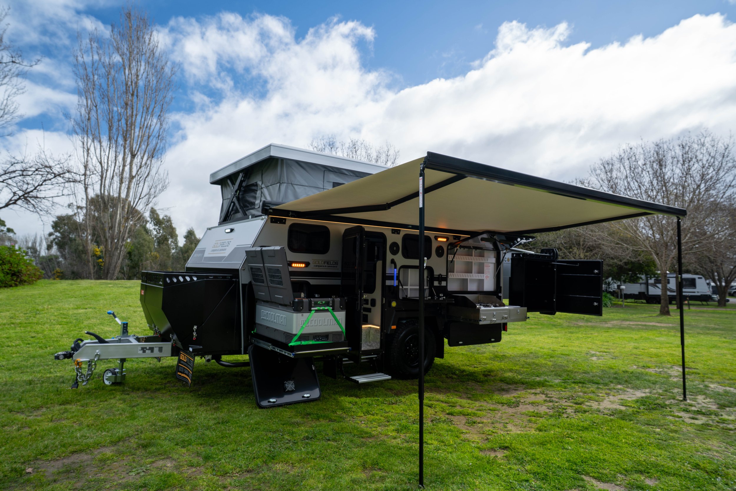 A recreational vehicle with an extended awning set up on a grassy area, with trees and a partly cloudy sky in the background.
