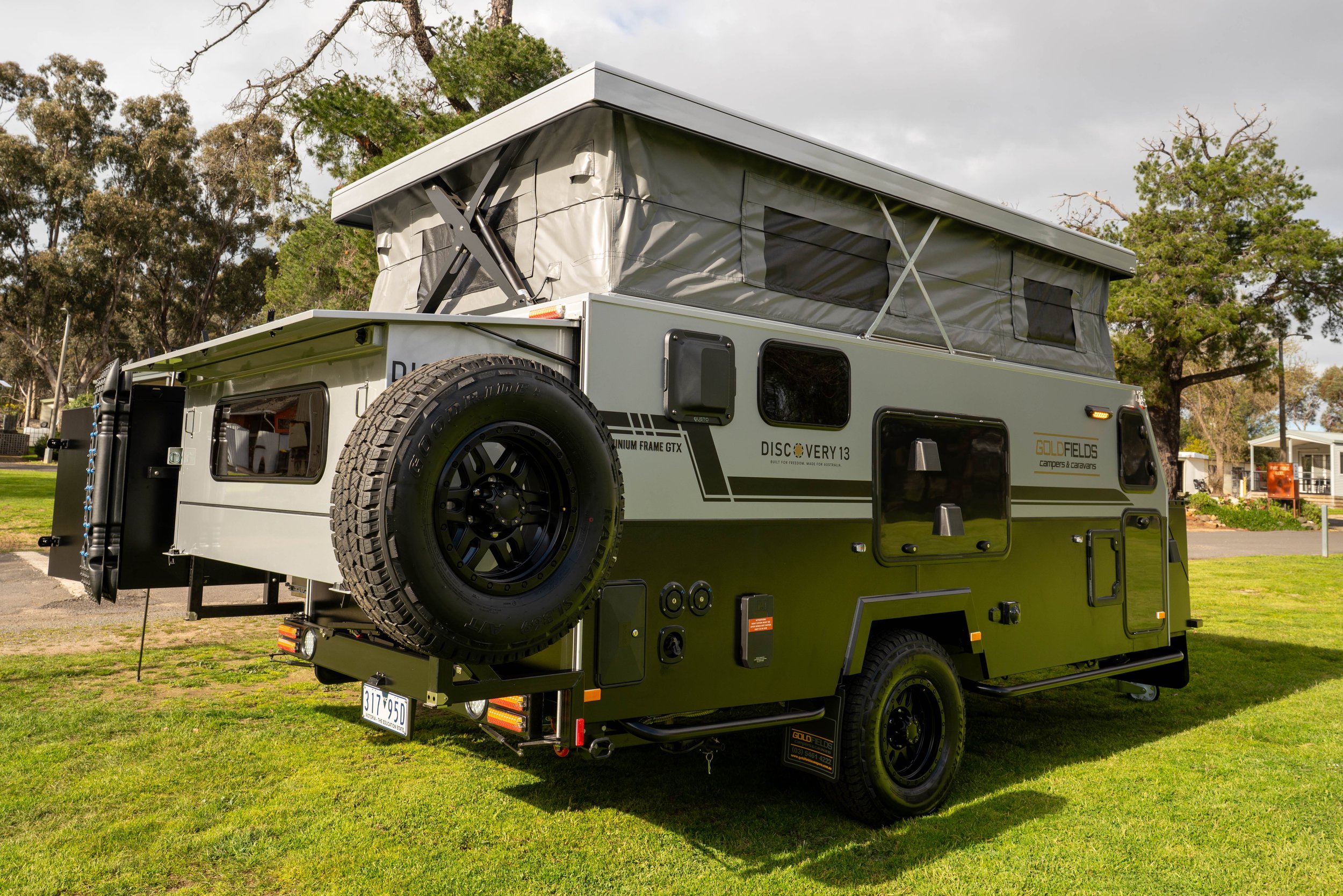 A compact camper trailer with a pop-up roof tent parked on grass, surrounded by trees and a cloudy sky.