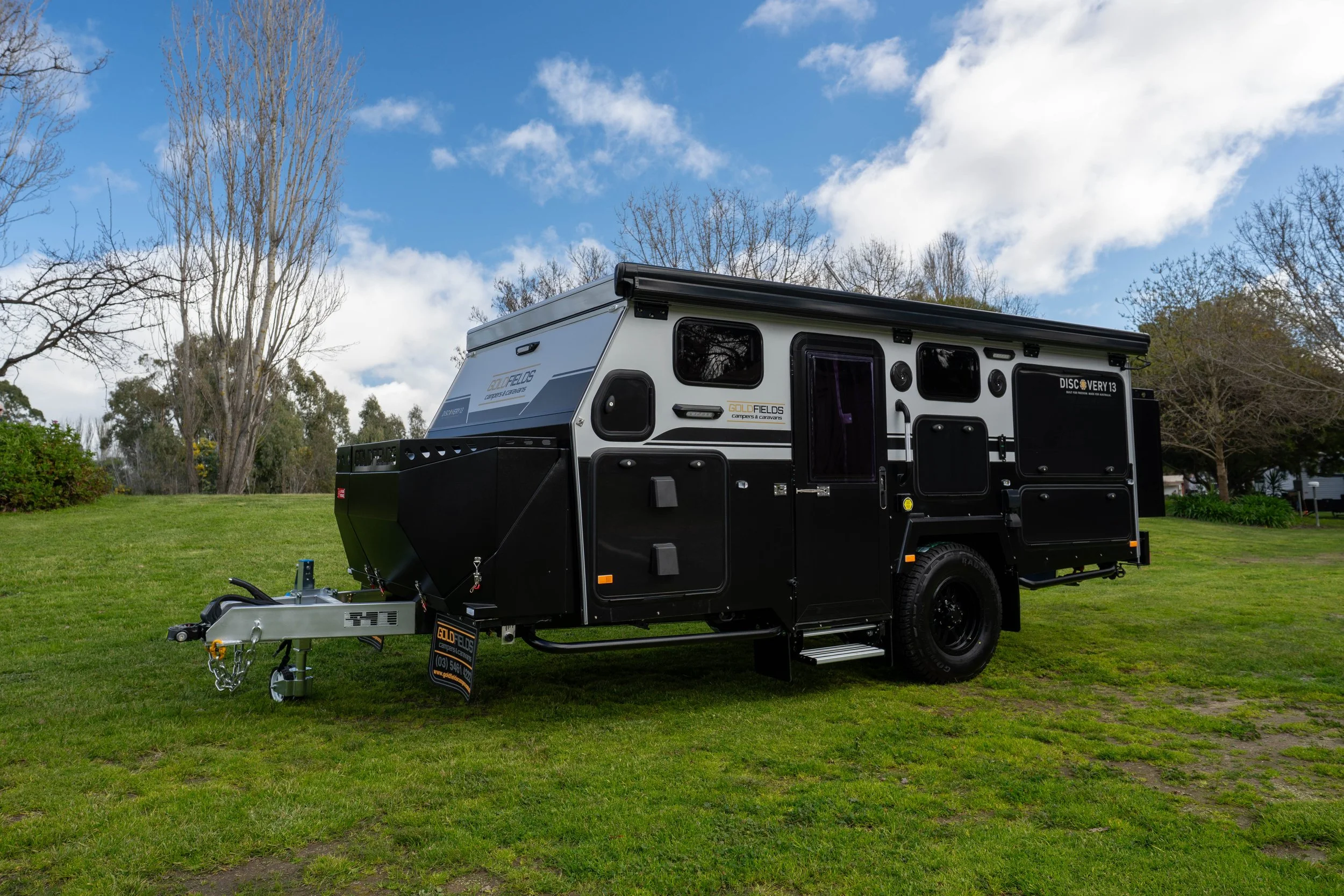 A black and grey camper trailer with outdoor storage compartments, set on a grassy area with trees and partly cloudy sky in the background.