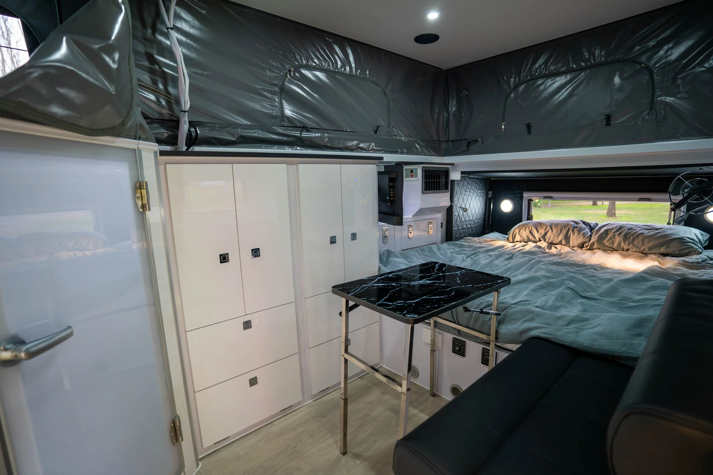 Interior of a camper van or tiny home showing a bed, white cabinets, a small black marble table, and a window with a view of trees outside.
