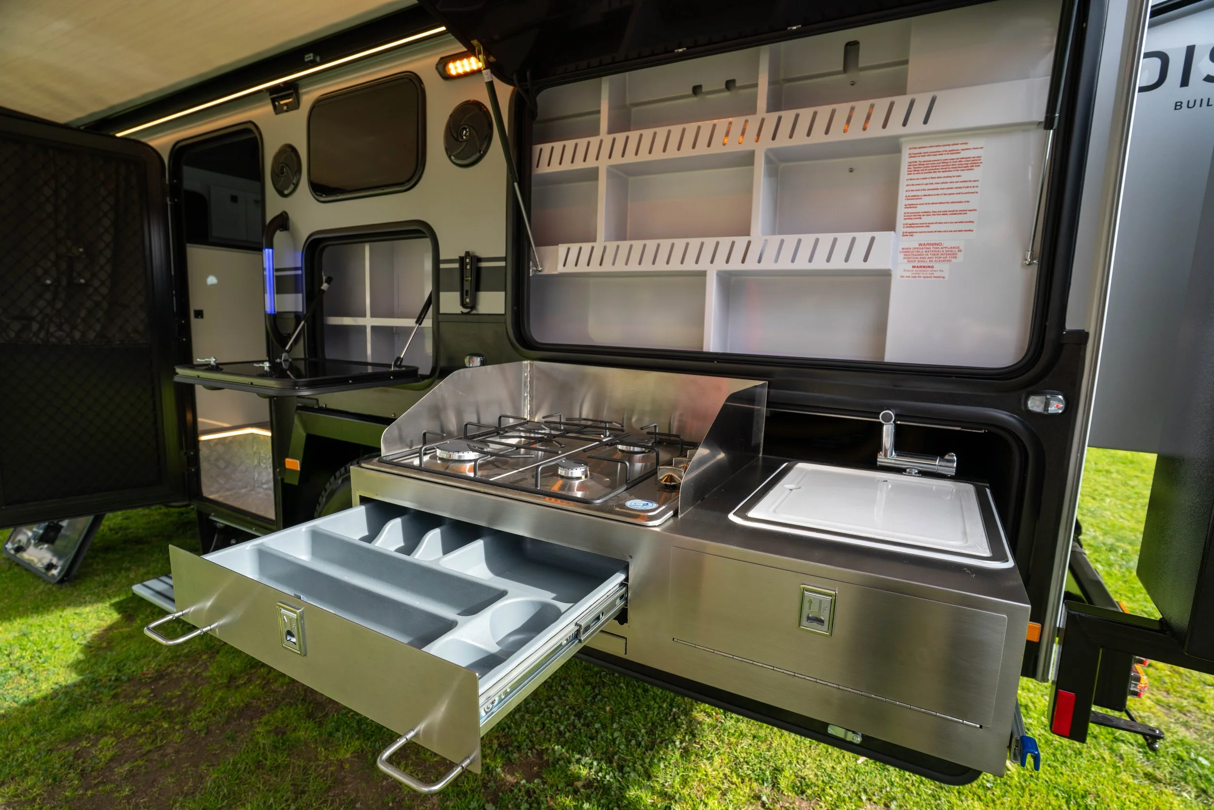View of a compact outdoor kitchen setup inside a trailer, featuring a stovetop, multiple storage drawers, and several open shelves.