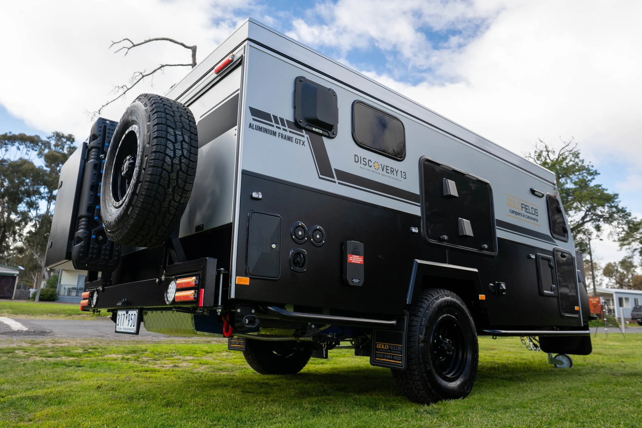 A modern camper trailer with large tires, mounted on a black chassis, parked on a grassy area with trees and buildings in the background, under partly cloudy skies.