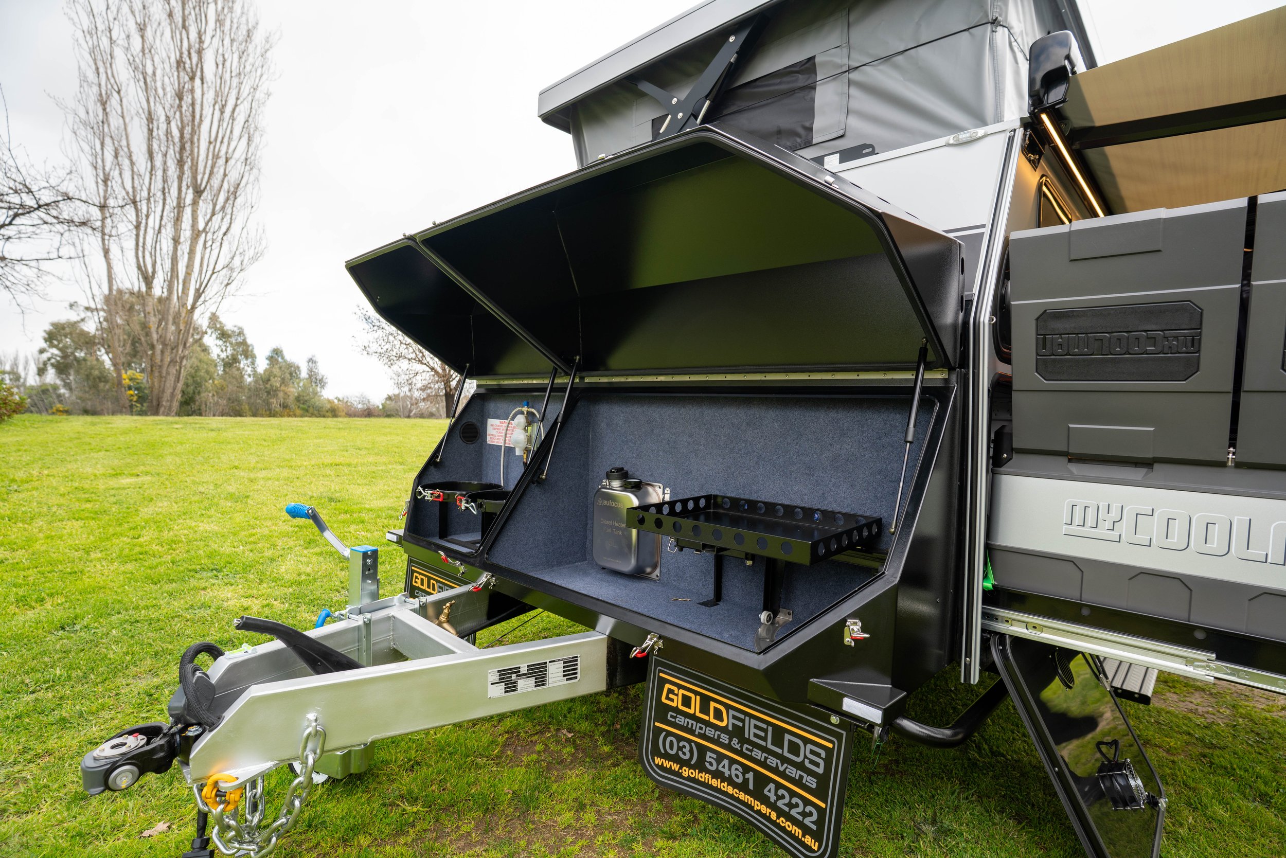 A portable camper kitchen setup on a trailer with a fold-out workspace, storage compartments, and a sign advertising Goldfields campers and caravans, situated on a grassy field with trees in the background.