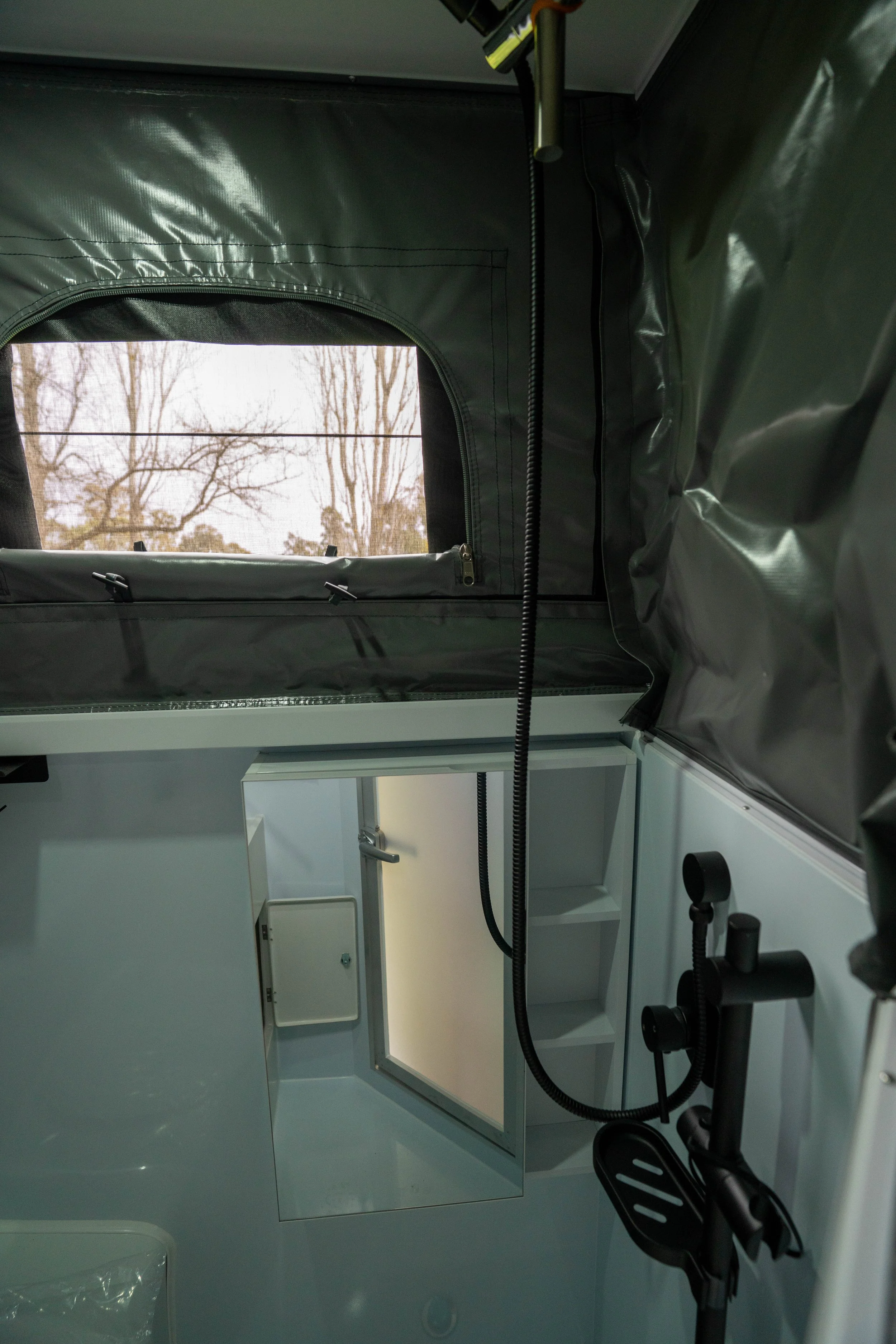 Interior of a medical or laboratory room with a large window showing leafless trees outside, and a white cabinet with a small mirror or glass panel, along with some black or dark-colored equipment, including a flexible hose and foot pedals.