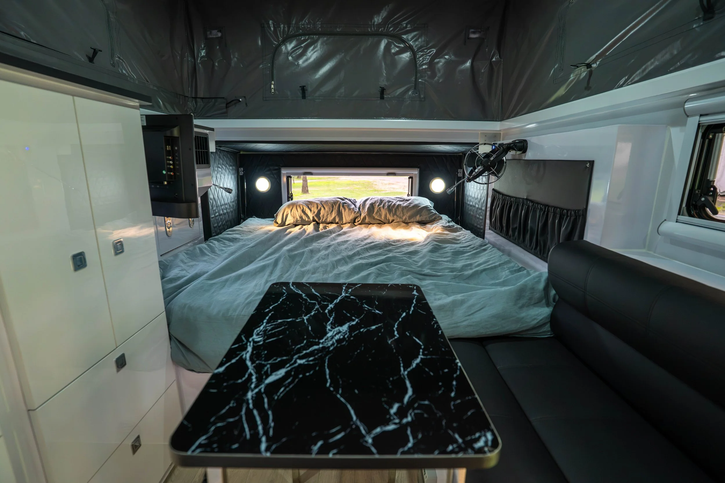 Interior of a modern camper with a bed, black marble-patterned table, and built-in storage cabinets, with windows and a view outside.