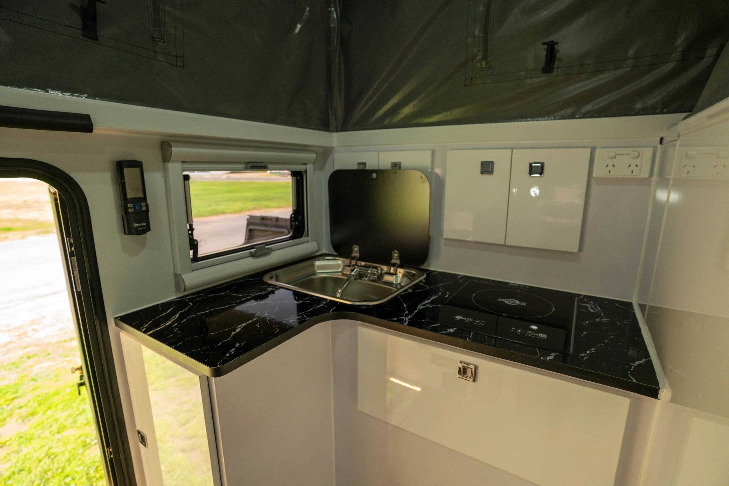 Compact kitchen space inside a camper van with a black marble countertop, small sink, window, and several white cabinets and electrical outlets on the wall.