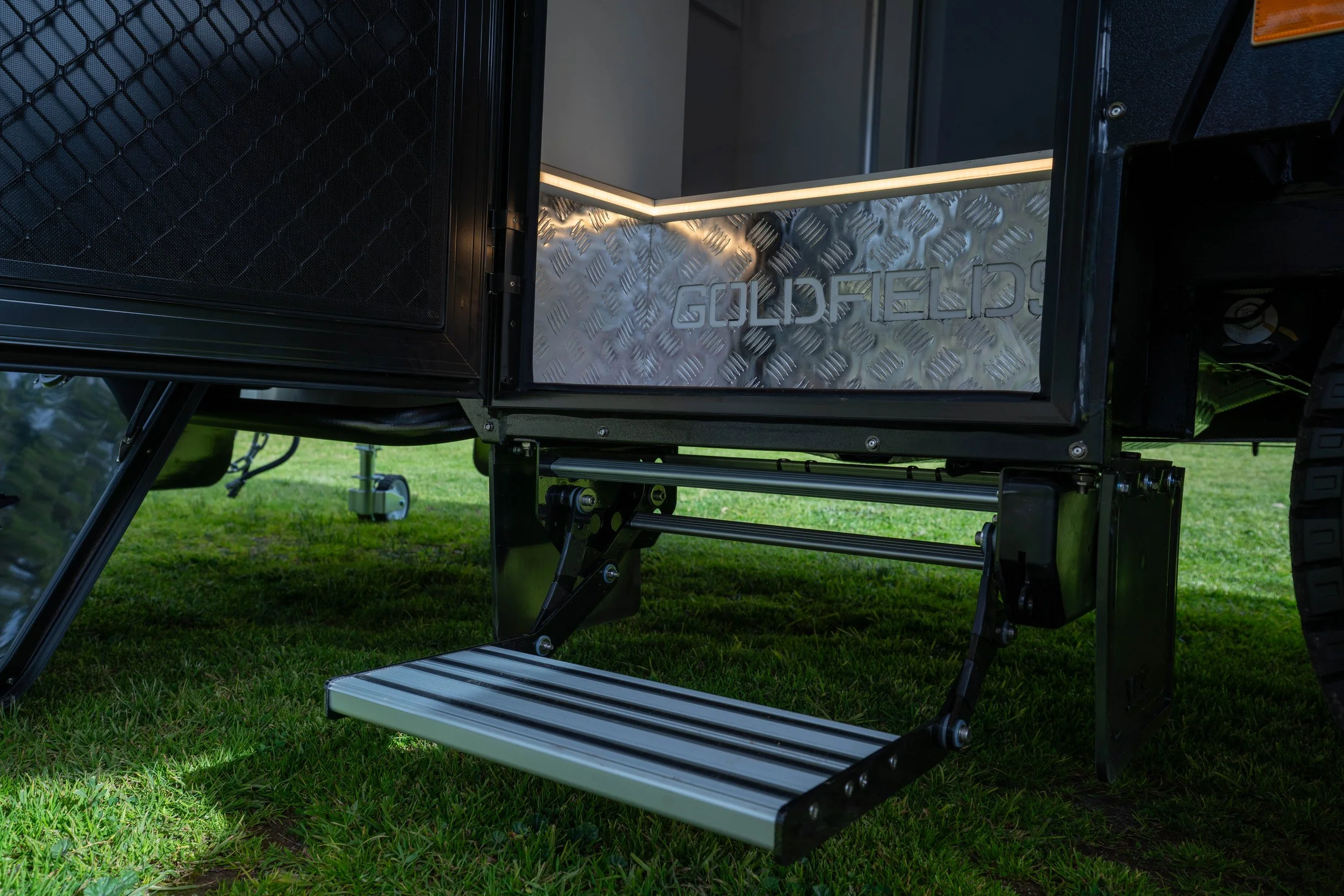 Close-up of a mobile food trailer with a metallic exterior, black mesh doors, and illuminated sign that reads 'GOLDFED'. The trailer is on a grassy outdoor setting.