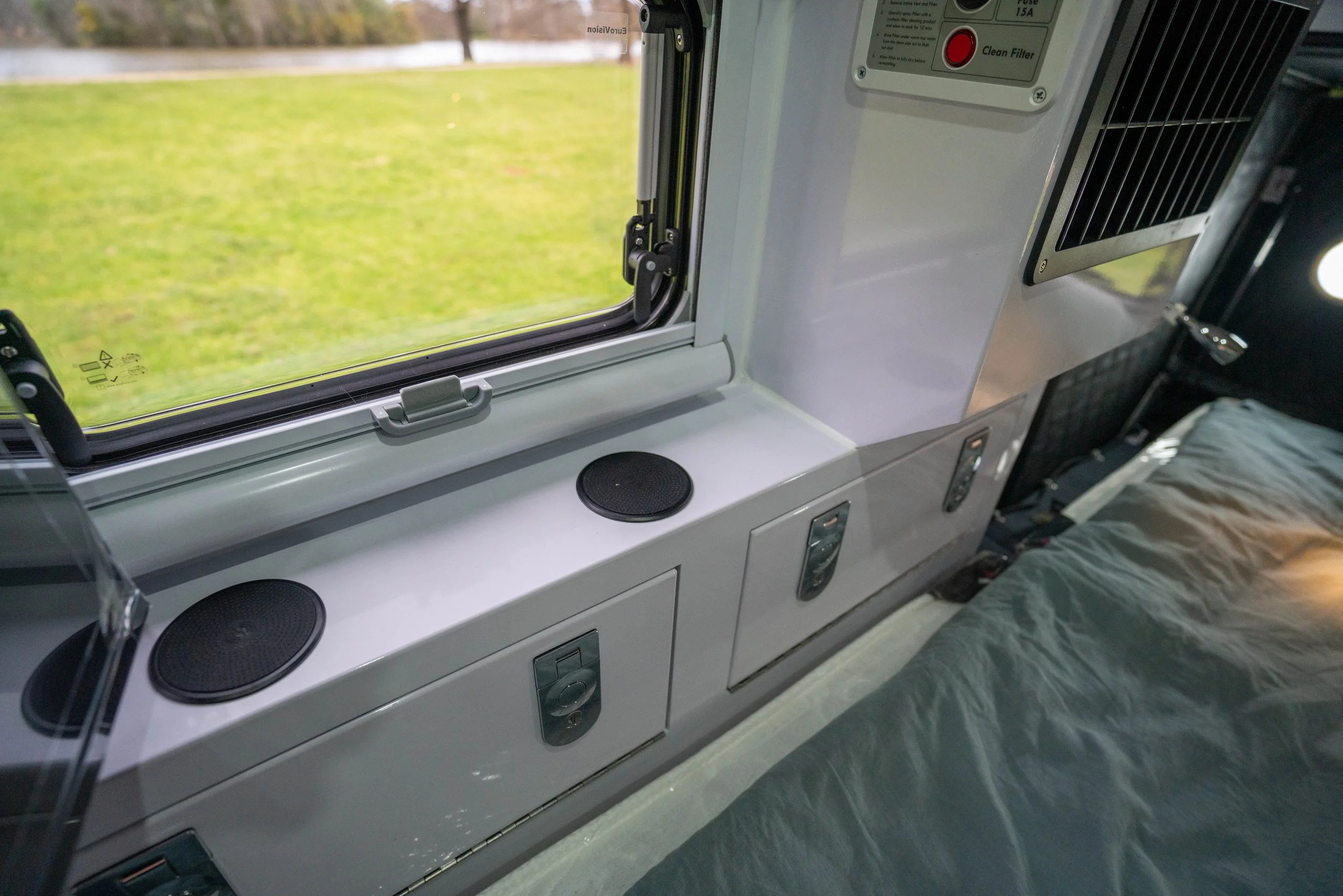 Interior of a recreational vehicle with a window showing a grassy outdoor landscape; includes a countertop with built-in speakers and a control panel.