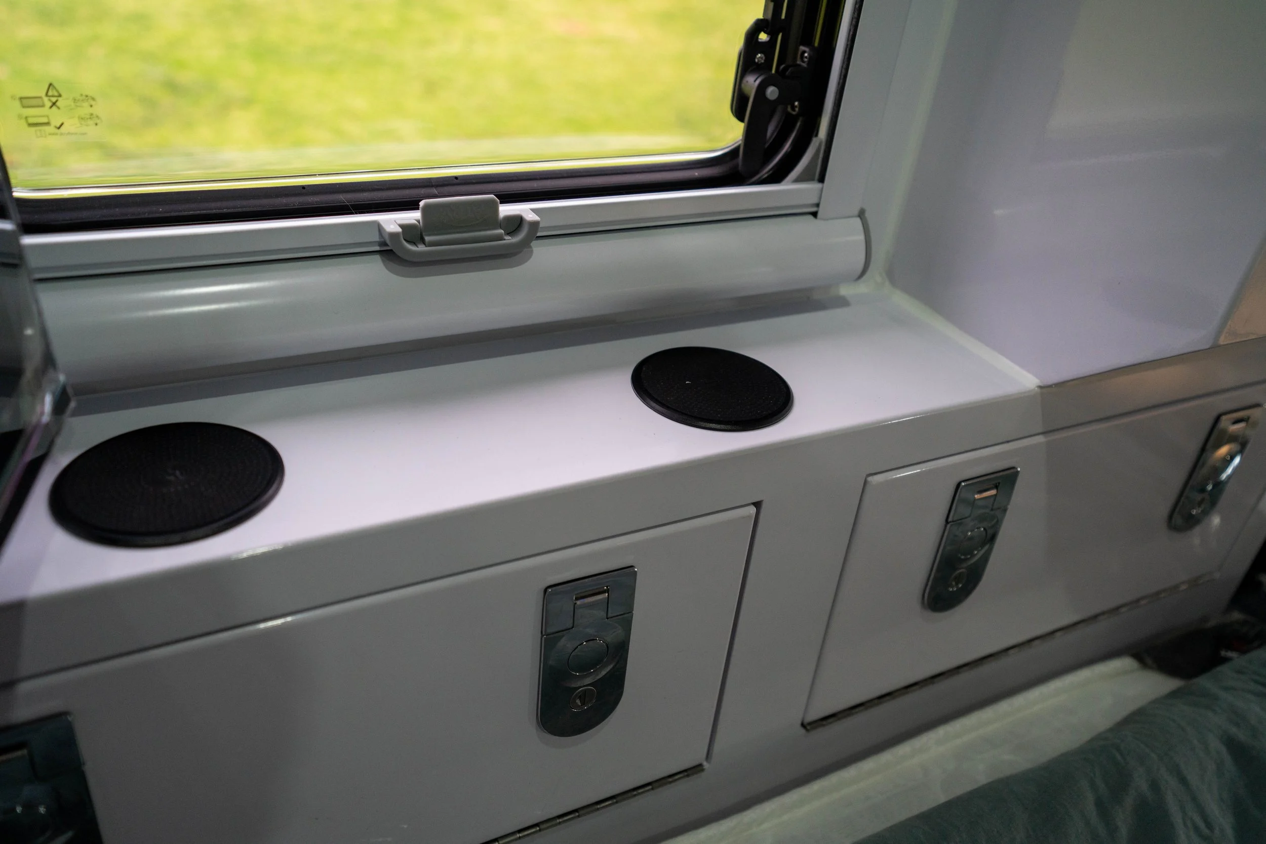 View of a window in an airplane or train with a handle latch and a yellow-green blurred landscape outside, and a white shelf below the window with black round speakers and metal latches.