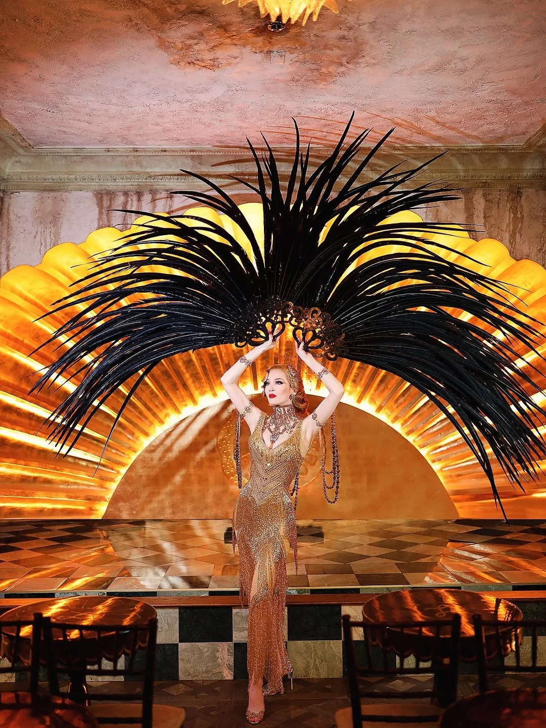 A woman in an ornate gold dress with jewelry poses on a stage with a large black decorative piece above her, illuminated by warm orange lighting.