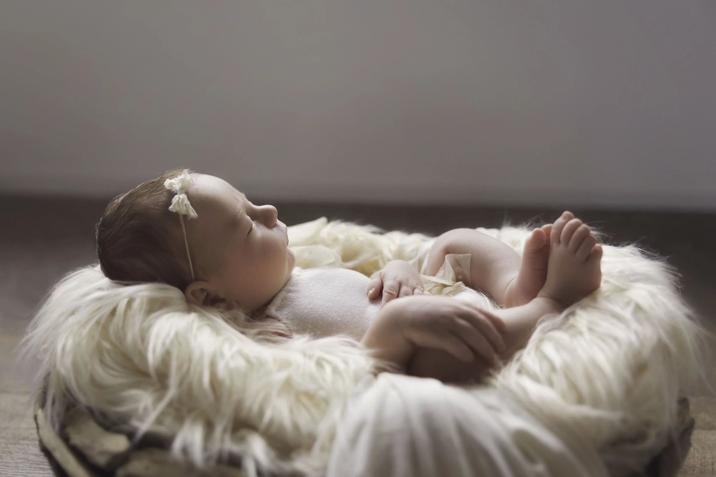 A newborn girl asleep on cream fluffy blanket in a wooden basket with a sweet little cream bow and soft knitted cream onesie
