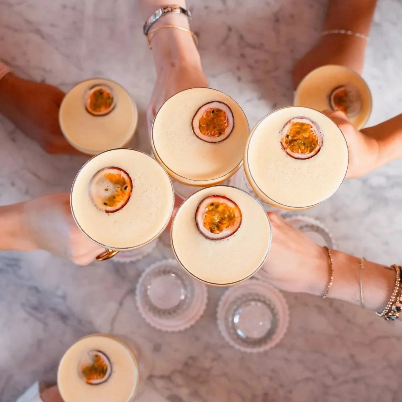 Syv personer som skåler med alkoholholdige cocktailer med pasjonsfrukt pyntet på toppen, sett ovenfra over en hvit marmortabell.