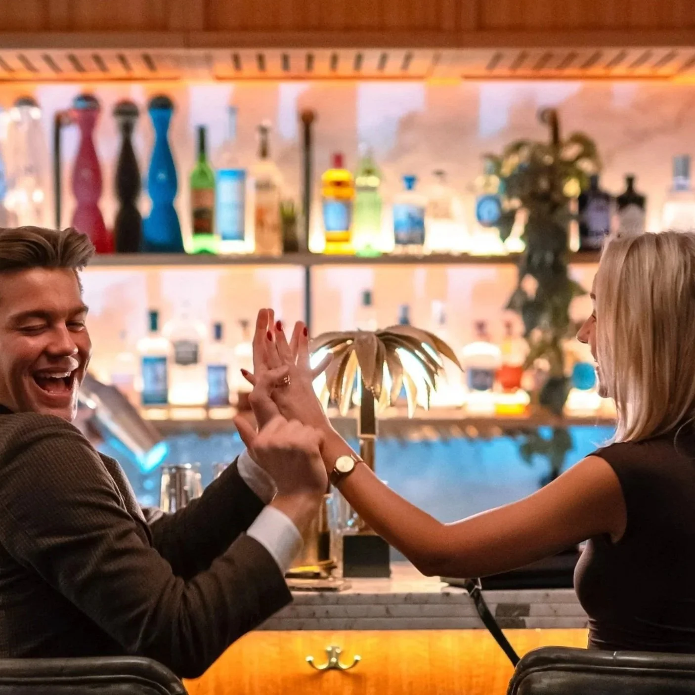 To personer på en bar som håndhilser og ler, med hyller fulle av alkoholflasker i bakgrunnen.