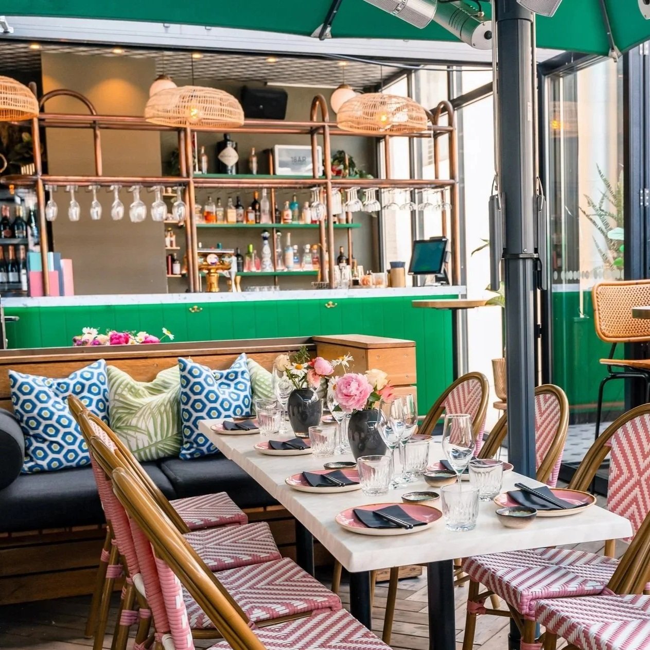 Innendørs restaurant med borddekket for gjester, blomsterdekorasjoner, komfortable stoler, og en bar i bakgrunnen med speil og hengeglass.