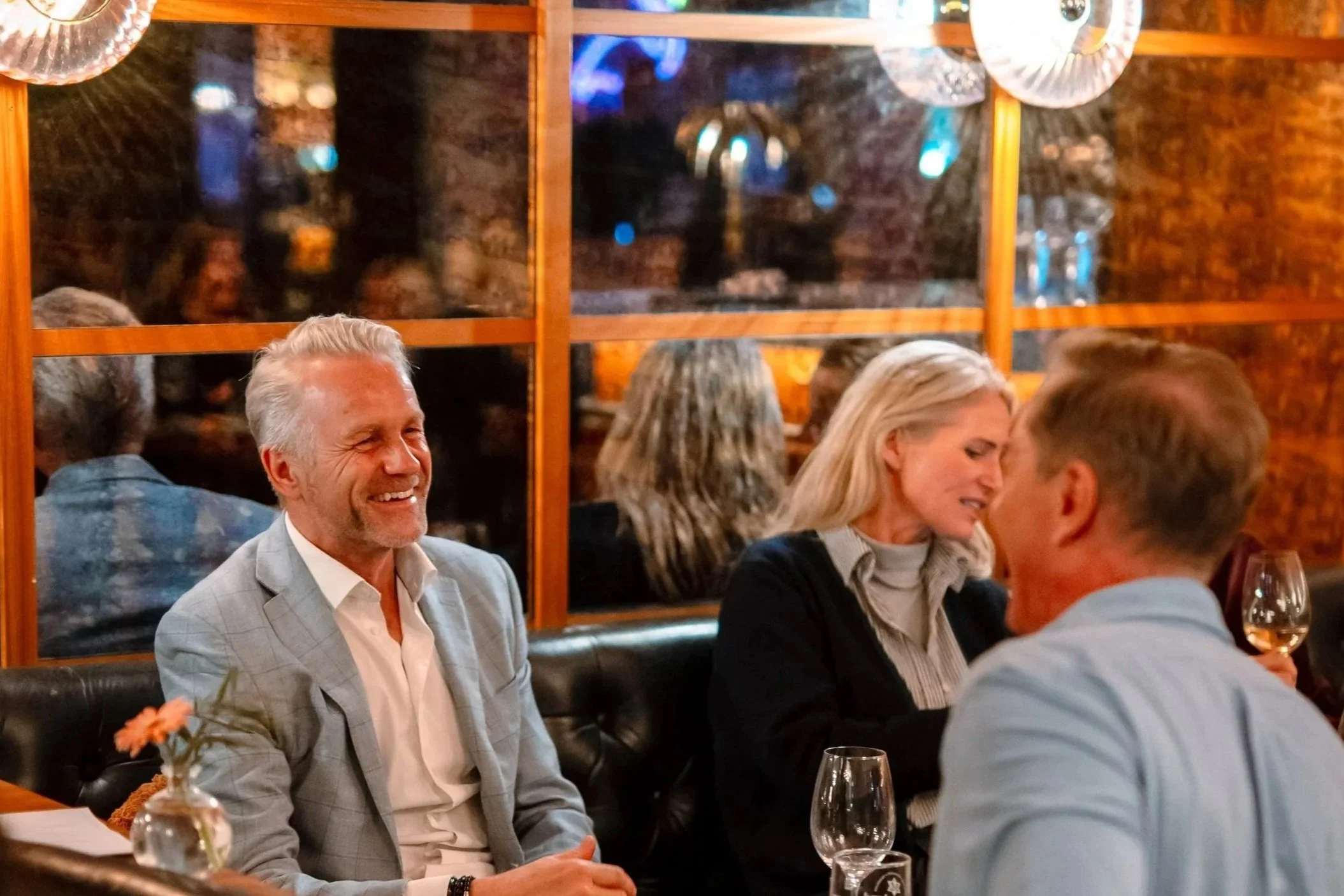 Menn og kvinner ler og prater på restaurant med vin og drinker, lyst og trivelig omgivelser.