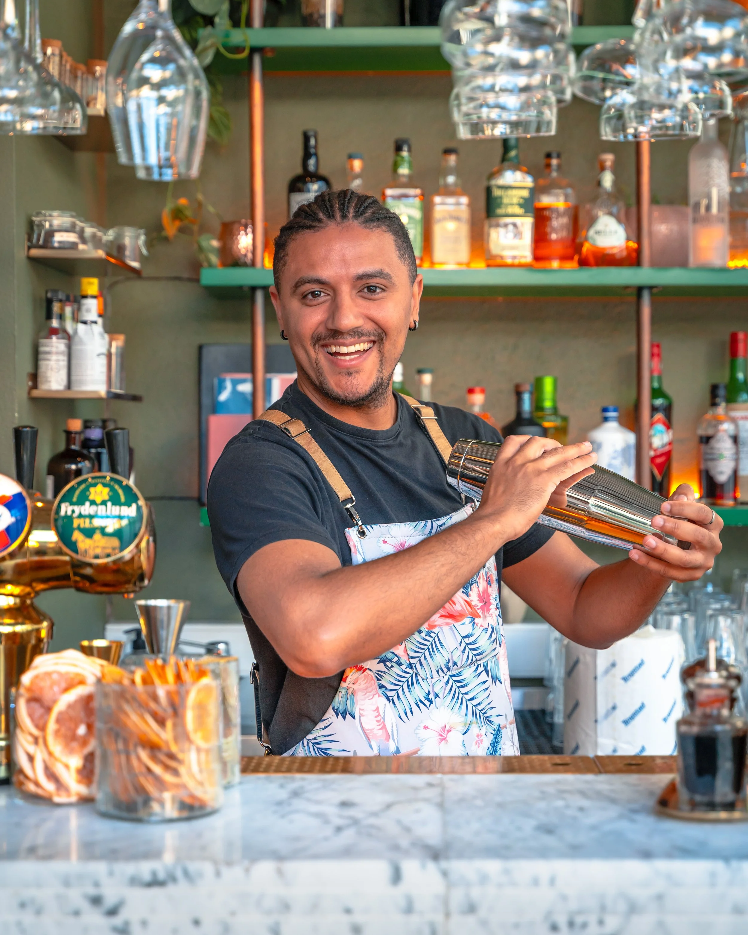 En smilende bartender som shakerer en drink bak en bar med ulike alkoholflasker og glass.