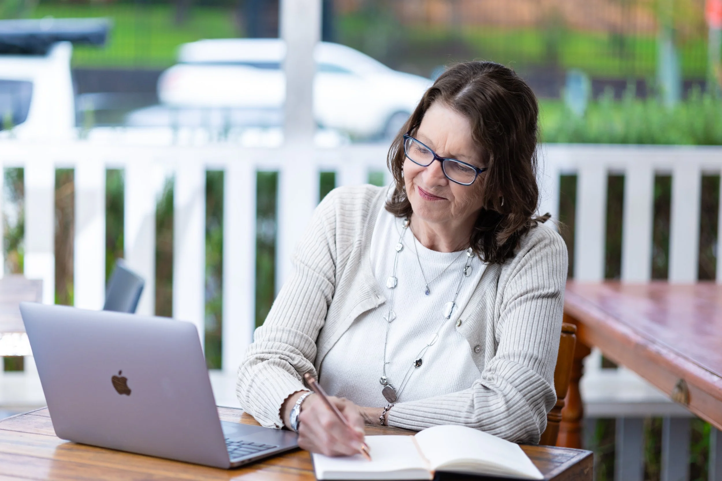 Woman with glasses sits at a wooden table outdoors, working on her laptop and writing in a notebook, with a white picket fence and greenery in the background.