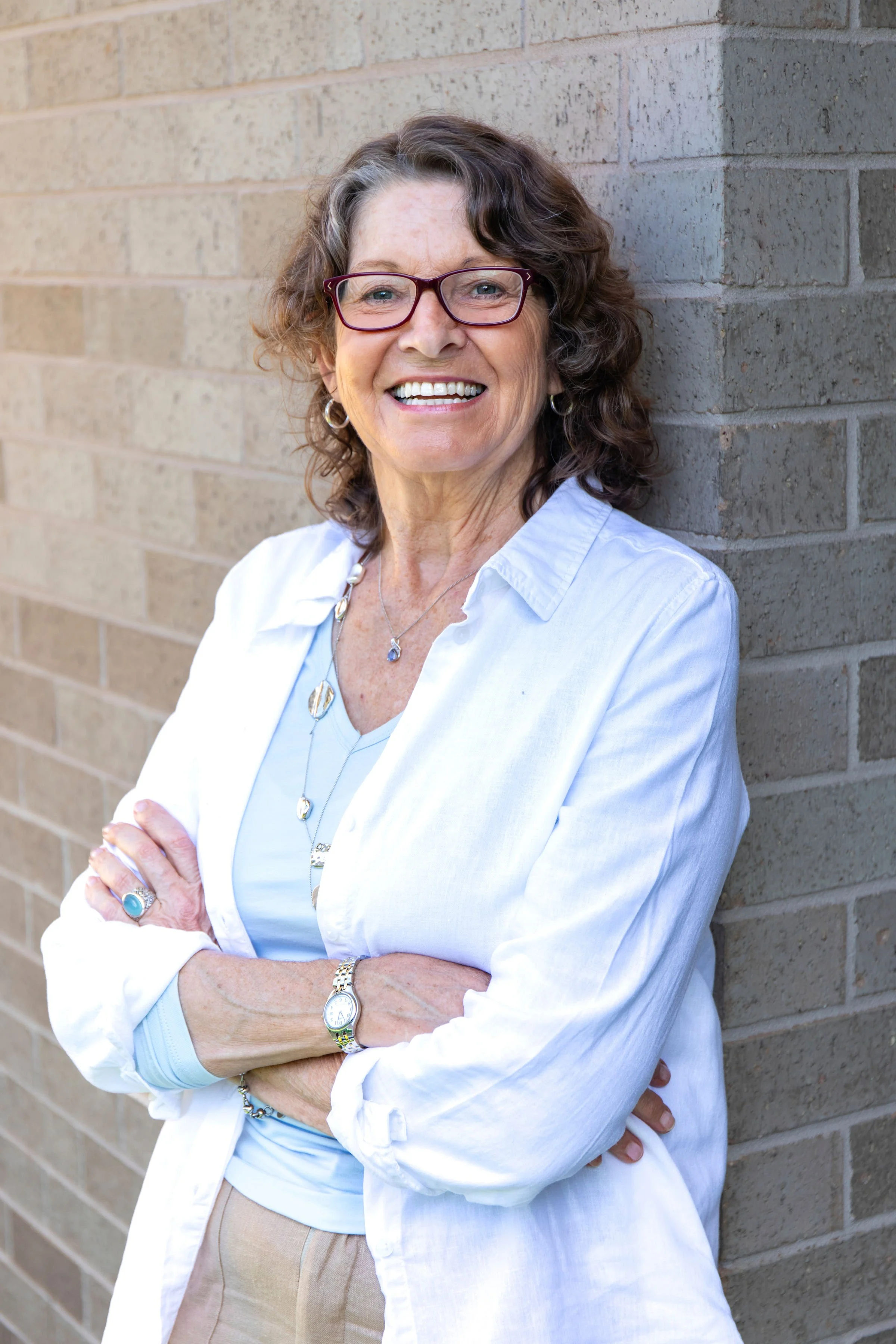 A smiling woman with curly brown hair and glasses standing with arms crossed in front of a brick wall.