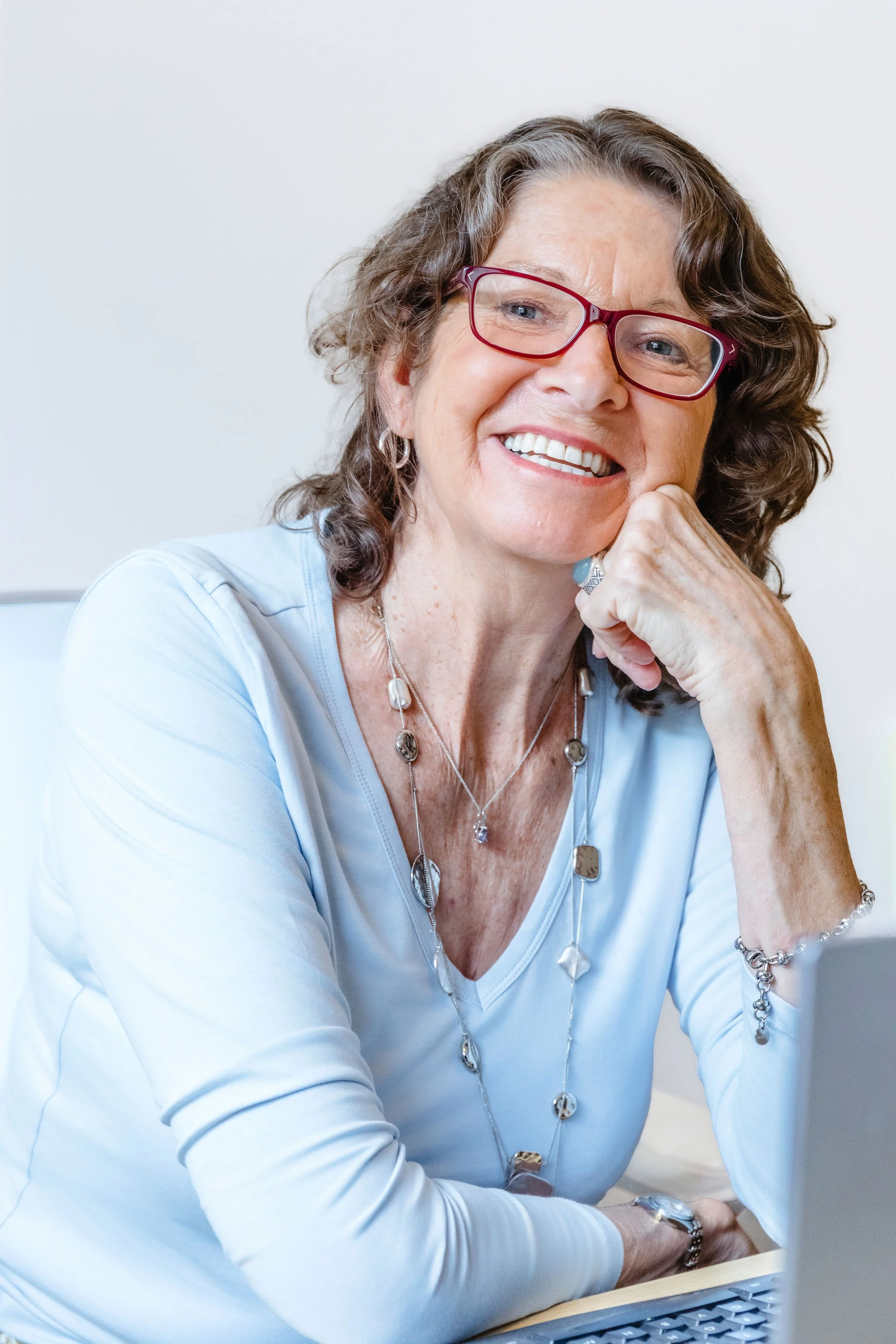 A smiling middle-aged woman with curly brown hair and red glasses wearing a light blue top, wearing jewelry, resting her chin on her hand, sitting at a desk with a keyboard.