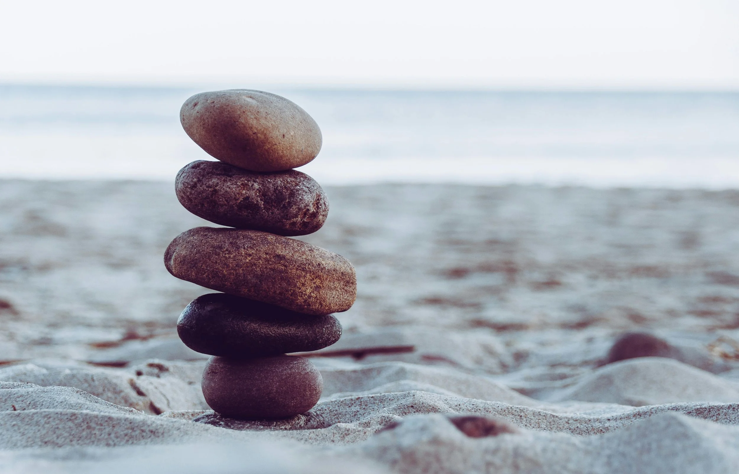 Five smooth stones stacked in a tower on a sandy beach with an ocean in the background.