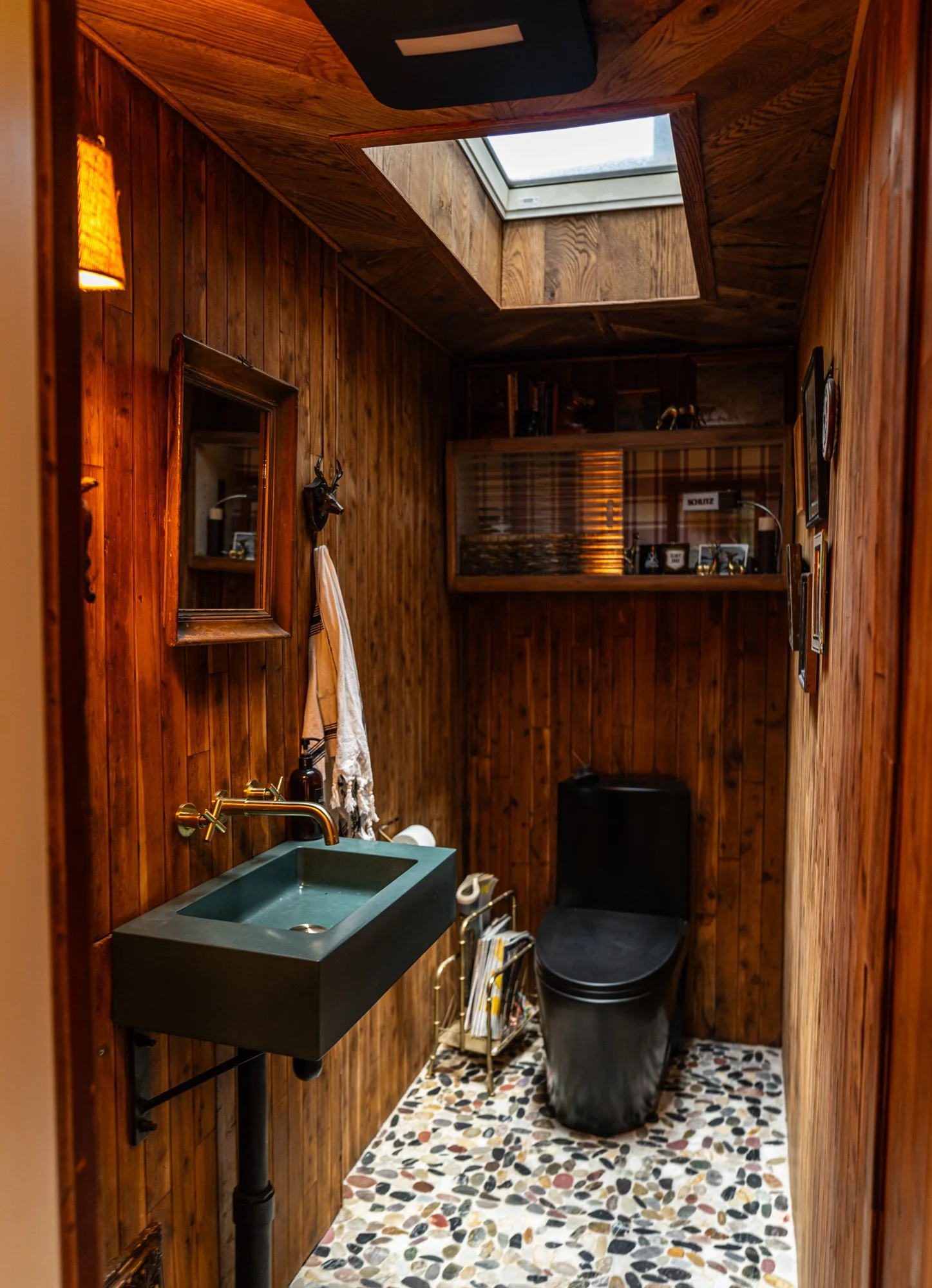 A small bathroom with wood-paneled walls and ceiling, a skylight, a black toilet, a small black sink, a mirror, a towel rack, and a colorful pebble-patterned floor.