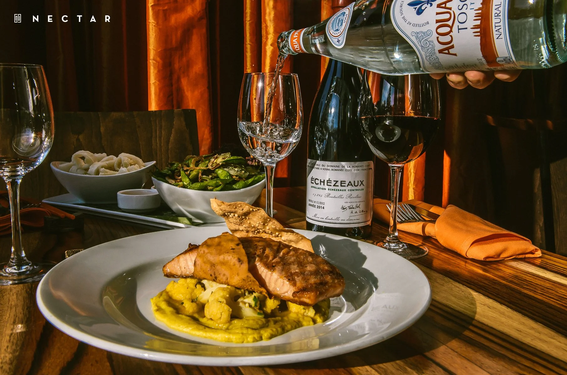 A gourmet meal featuring a plate with cooked fish on a bed of yellow mashed potatoes at a fine dining restaurant, with a glass of red wine, a bottle of sparkling water, and a salad in the background, along with other empty glasses and a cutlery set o