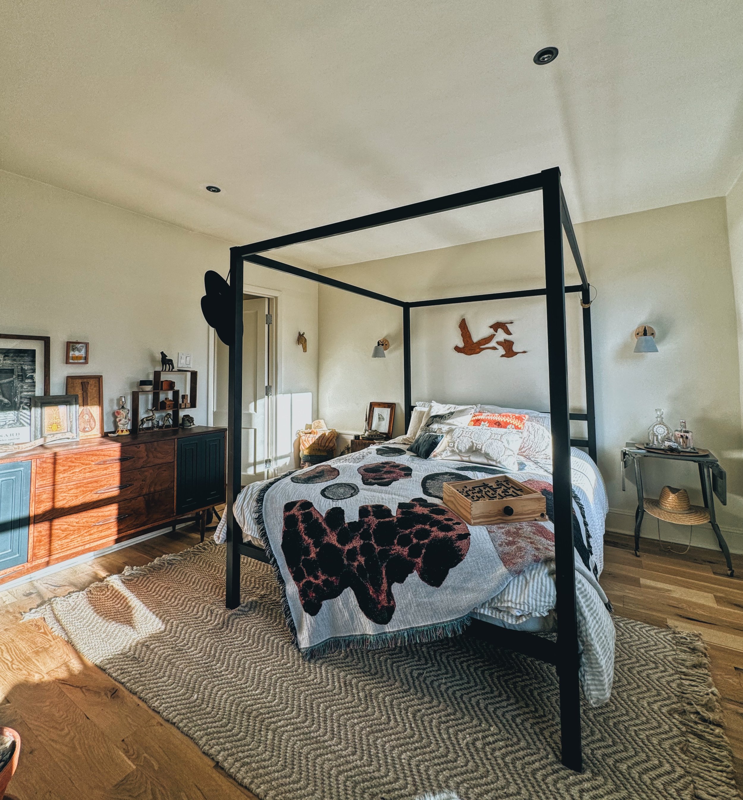 A bedroom with a black canopy bed, patterned bedding, wooden floors, a textured rug, a wooden sideboard with framed pictures and decorative items, and wall decorations including mounted animals and art pieces.