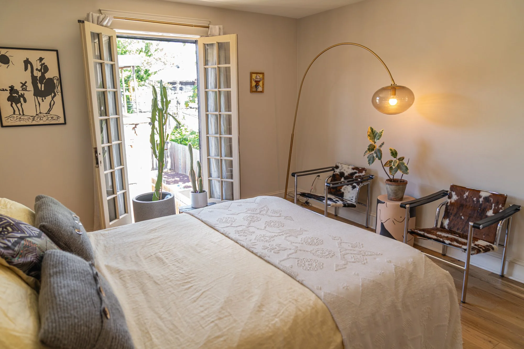 Cozy bedroom with a large bed, open French doors leading outside, and a cactus plant. The room features modern decor with vintage chairs, a leafy potted plant, a wall-mounted lamp, and artwork.