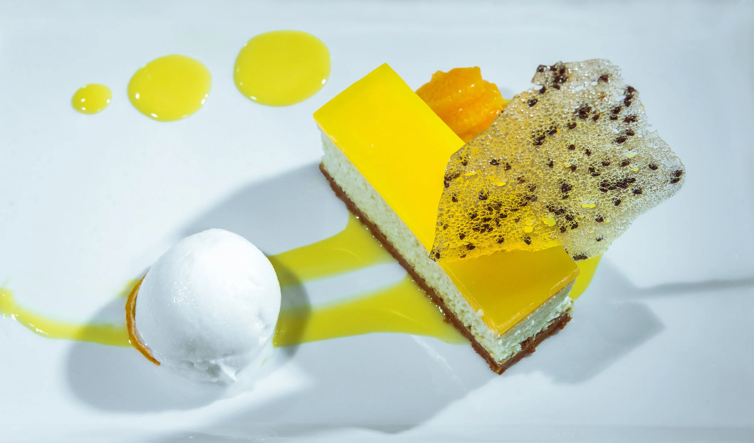 Close-up of a plated dessert featuring a layered cake with yellow glaze, a scoop of white ice cream, and decorative sauces and garnishes on a white plate.