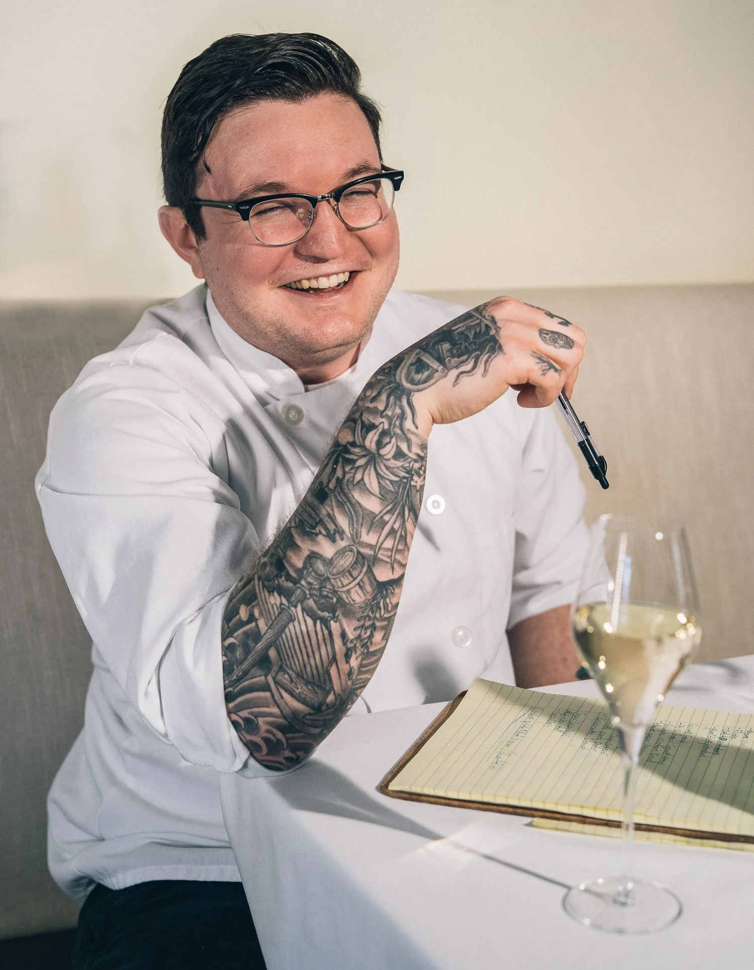 A smiling chef with glasses, tattoos on his arm, wearing a white chef coat, sitting at a table with a notebook and a glass of white wine.