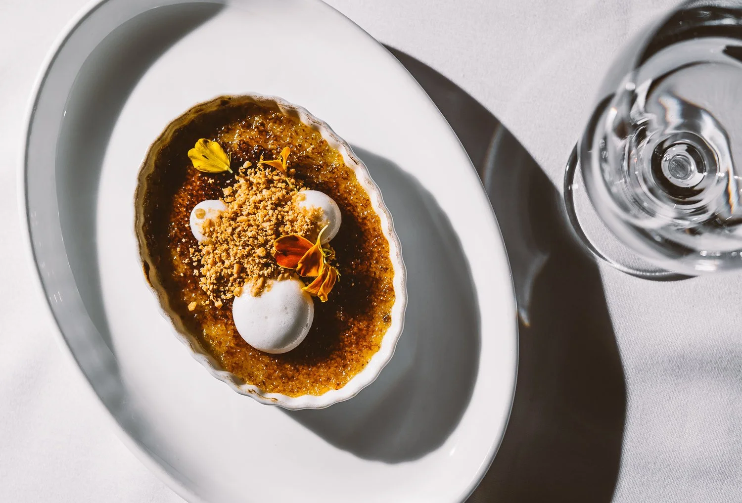 A dessert served in a white ramekin on a white plate, garnished with edible flowers and dollops of whipped cream, beside a glass of water.
