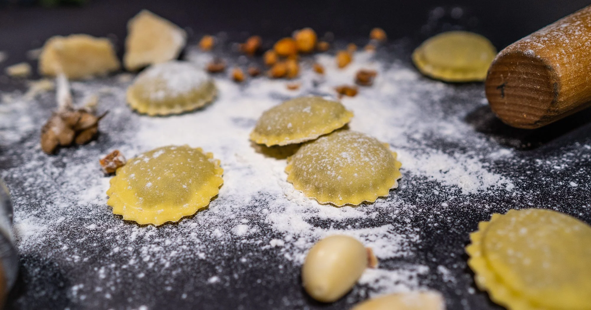 Unorganized homemade ravioli pasta on a floured dark surface, with scattered nuts, cheese pieces, and a wooden rolling pin.