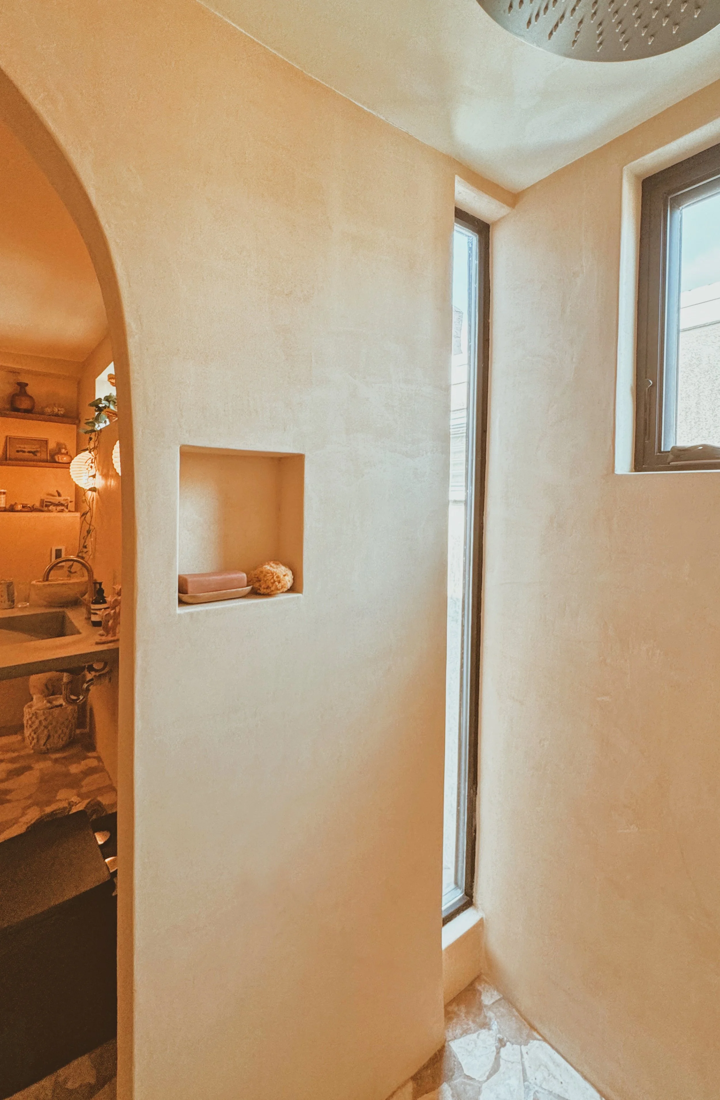 A narrow bathroom with beige textured walls, two small windows, and a built-in niche holding a soap dish and a decorative item. Part of a sink area with shelves and decorations is visible through an arched doorway.