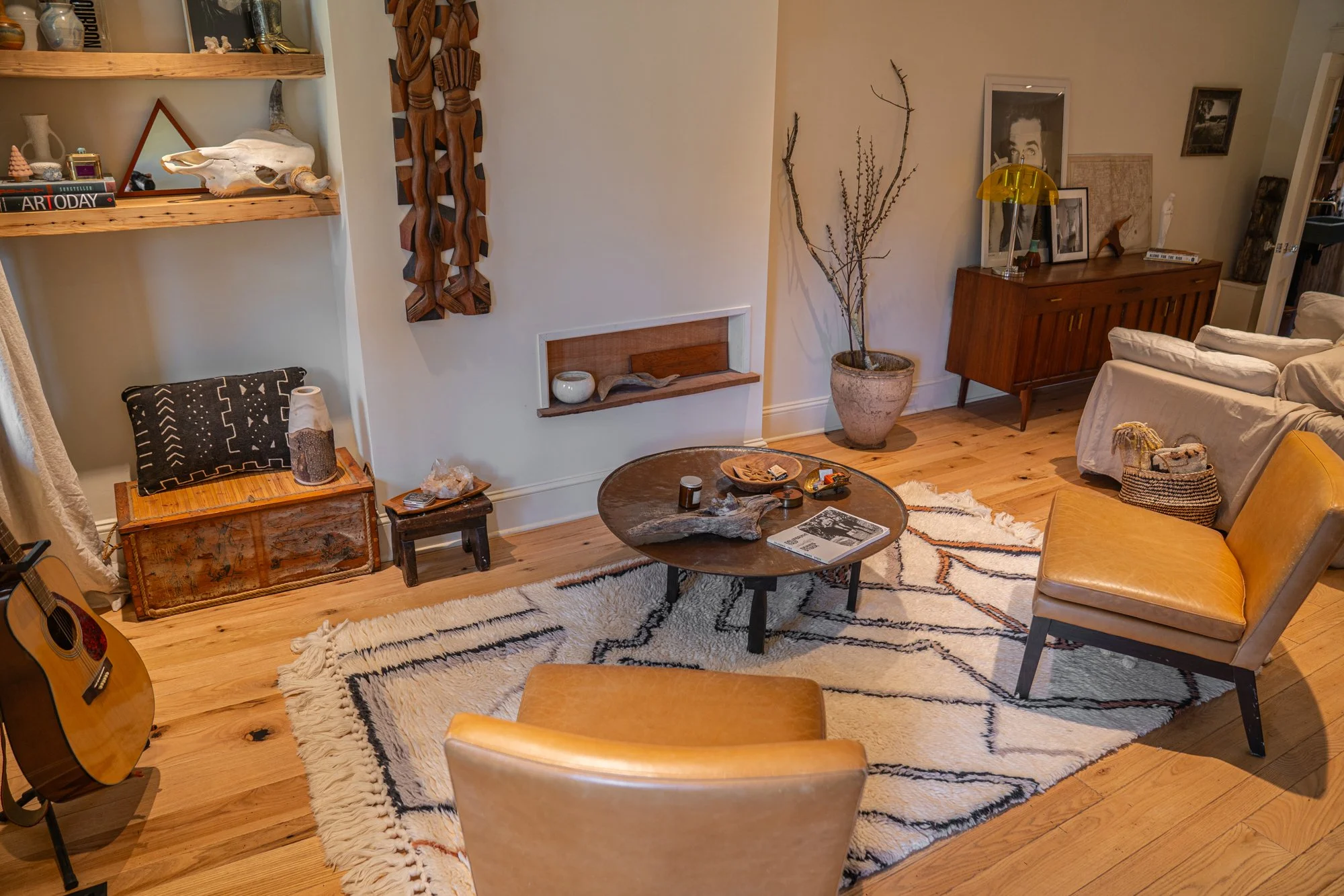 Living room with wooden floors, a patterned rug, a round wooden coffee table with decorative items, tan and beige chairs, a white sofa, a wooden sideboard with framed photos, a large potted plant, and a guitar.